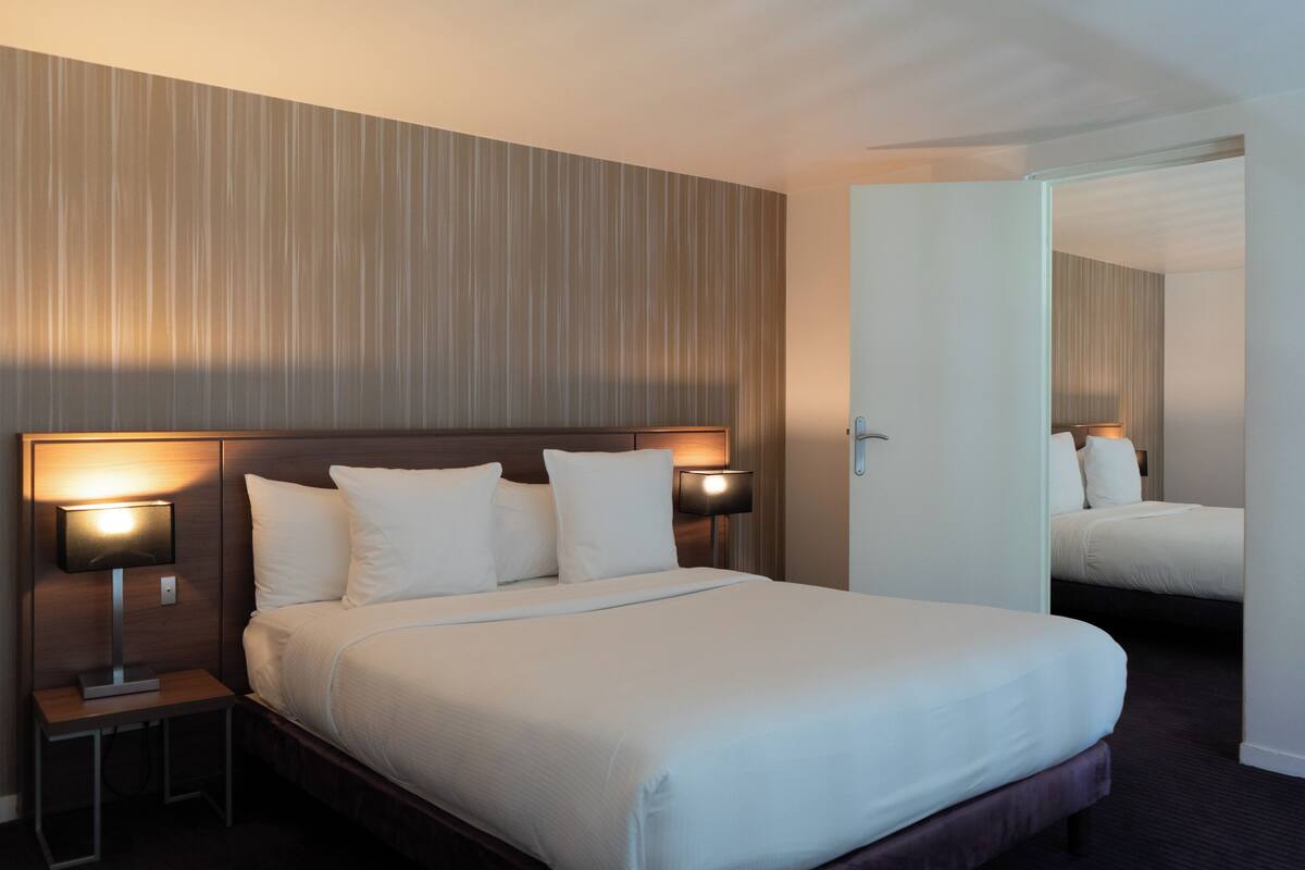 A hotel room with a large bed, two bedside tables with lamps, a mirror, and a door leading to another room.