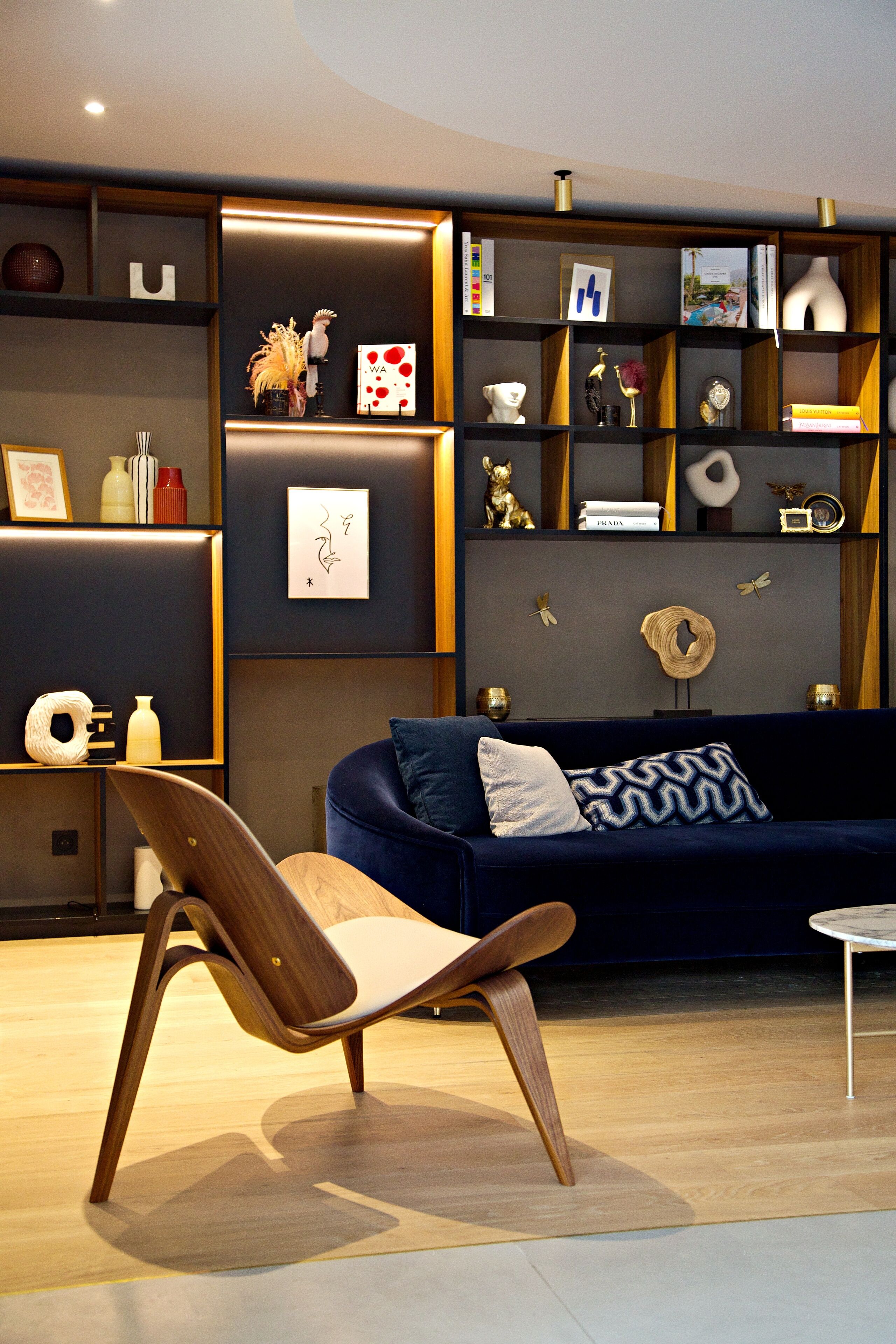 A modern living room with a dark blue sofa, a wooden chair, and a small round table. Shelves with decorative items and framed pictures are visible.