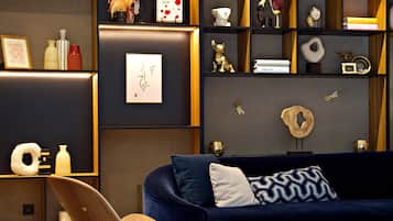 A modern living room with a dark blue sofa, a wooden chair, and a small round table. Shelves with decorative items and framed pictures are visible.