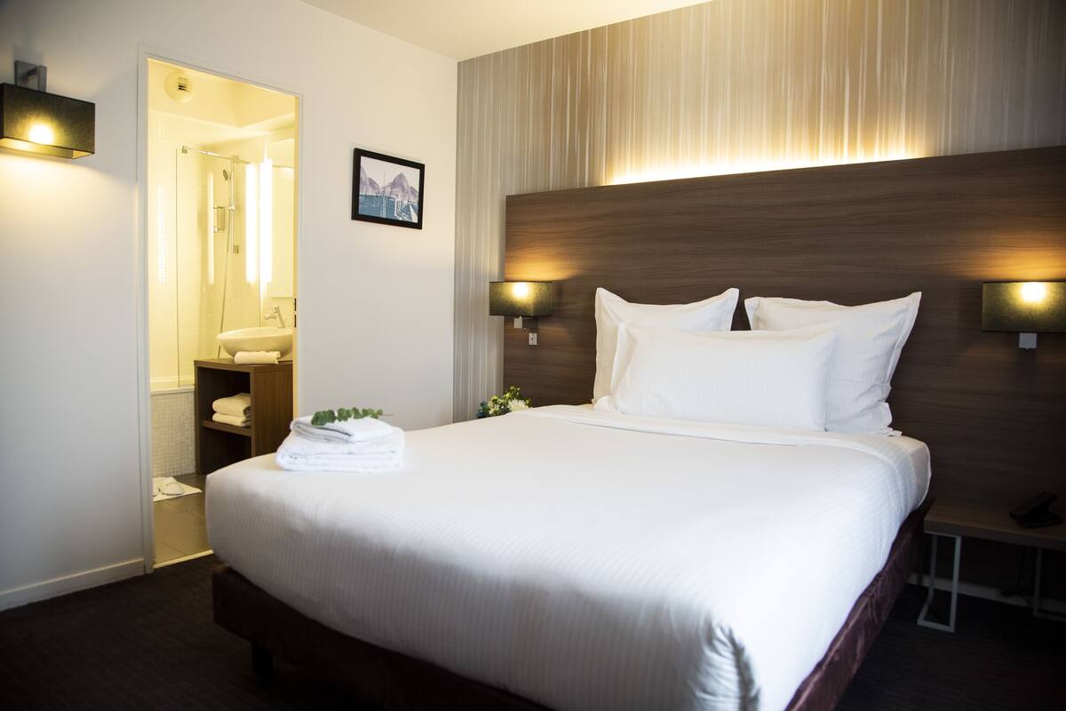 A hotel room with a large bed, a wooden headboard, two wall-mounted lamps, a bathroom with a glass shower enclosure, and a framed picture on the wall.
