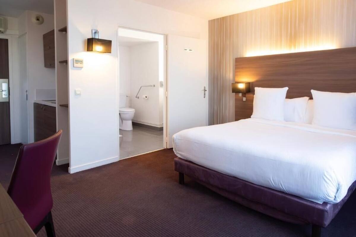 A hotel room with a large bed, a chair, a bathroom with a toilet and sink, and a wooden headboard with wall-mounted lights.