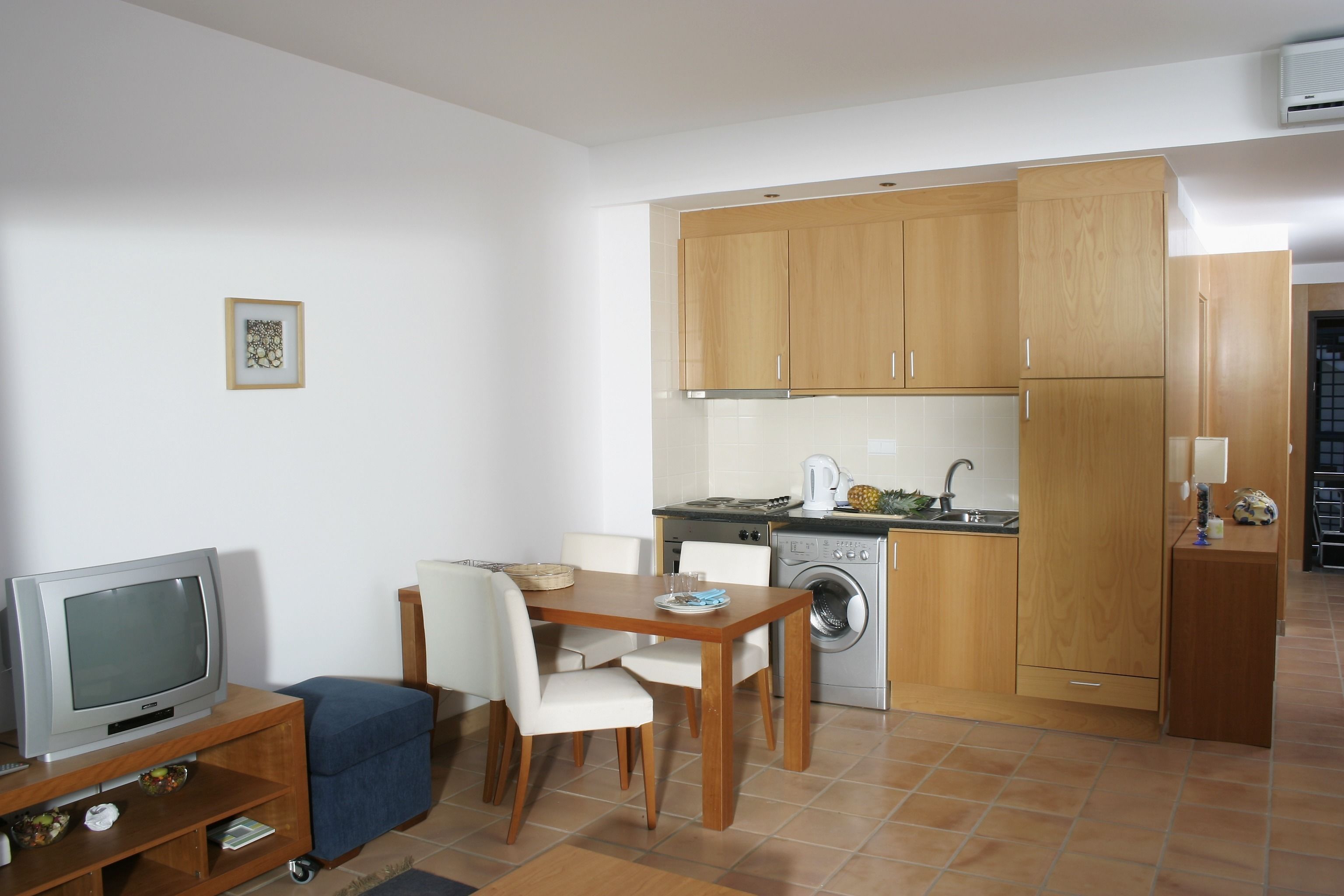 A compact kitchen with wooden cabinets, a washing machine, and a dining area with a TV and chairs.