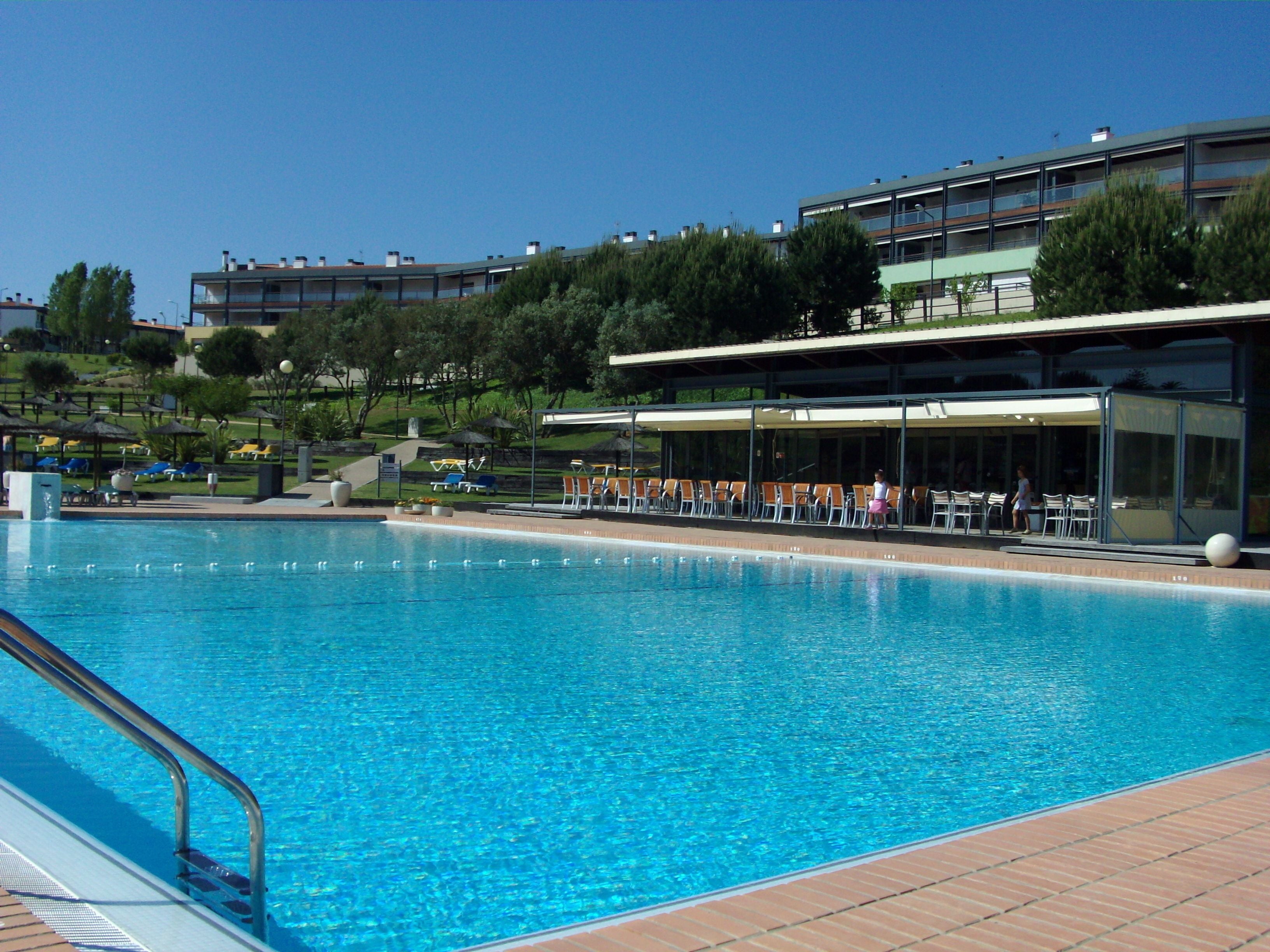 Una piscina con lettini e ombrelloni, circondata da edifici e alberi.