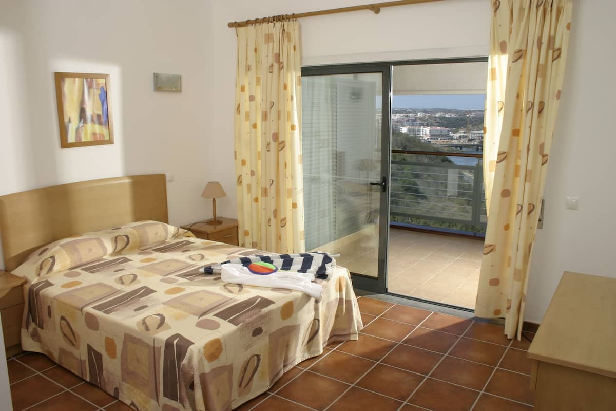 A bedroom with a bed, a nightstand, a lamp, a window with curtains, and a view of the city.