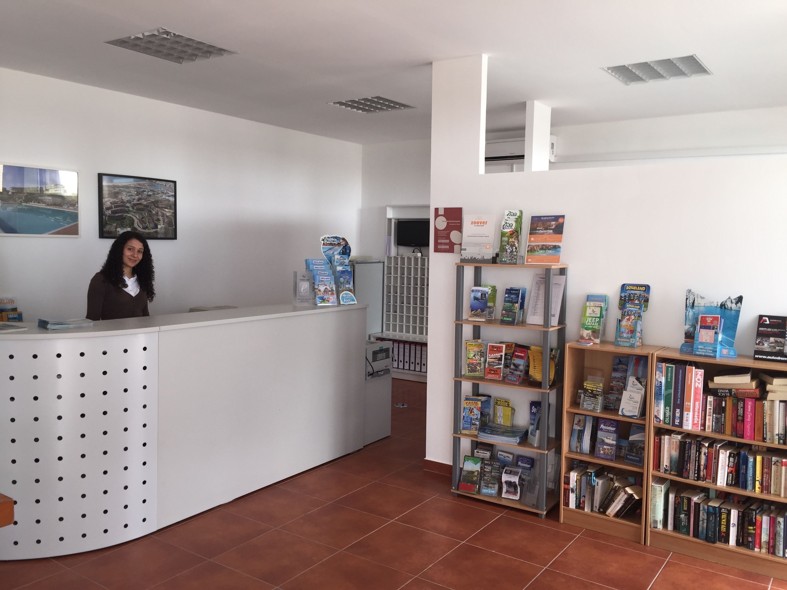 Zona de recepción con un mostrador blanco, una mujer de pie detrás y una estantería llena de libros de viajes.