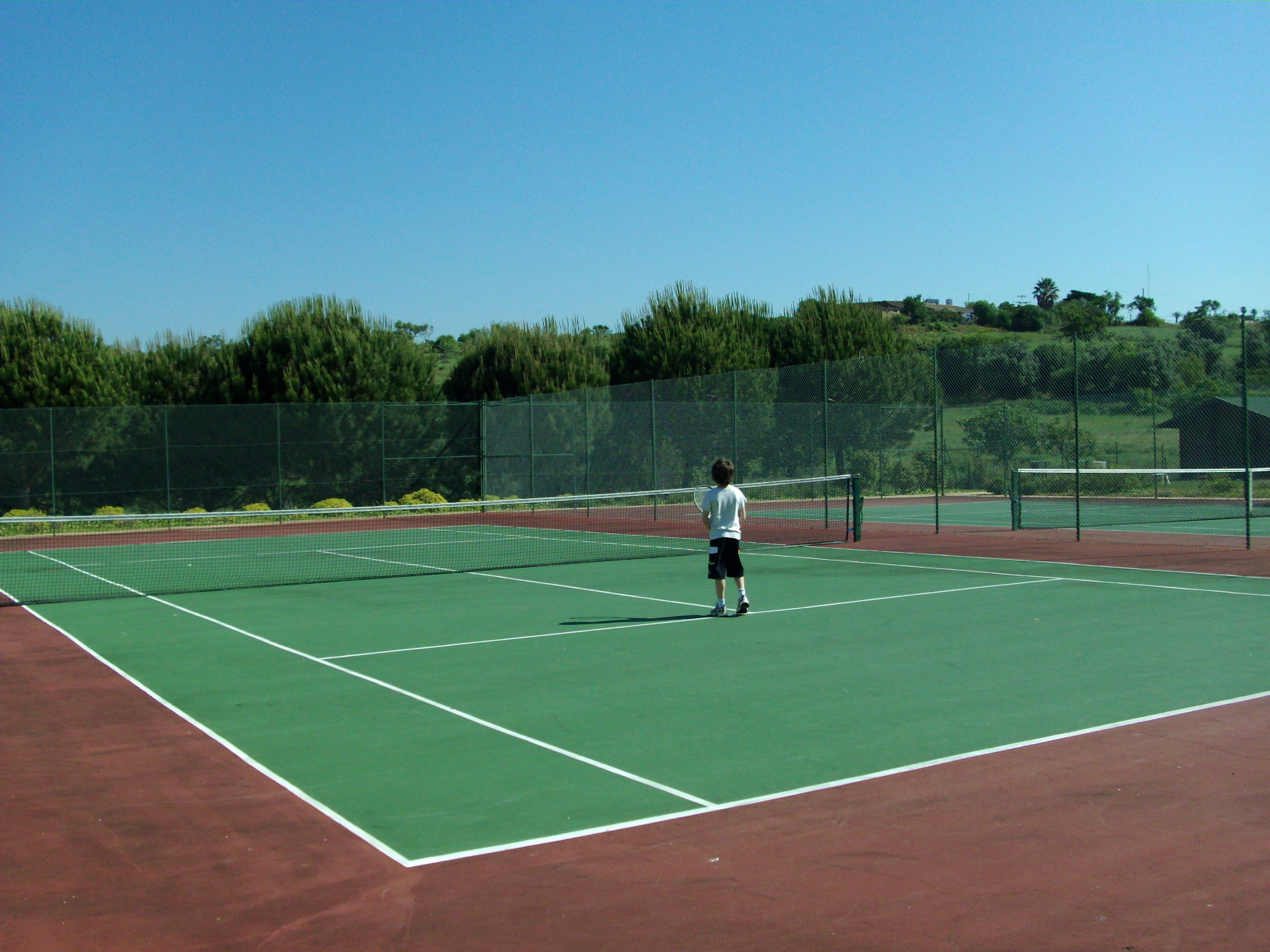 A tennis court with a net in the center, surrounded by a green fence and trees in the background.
