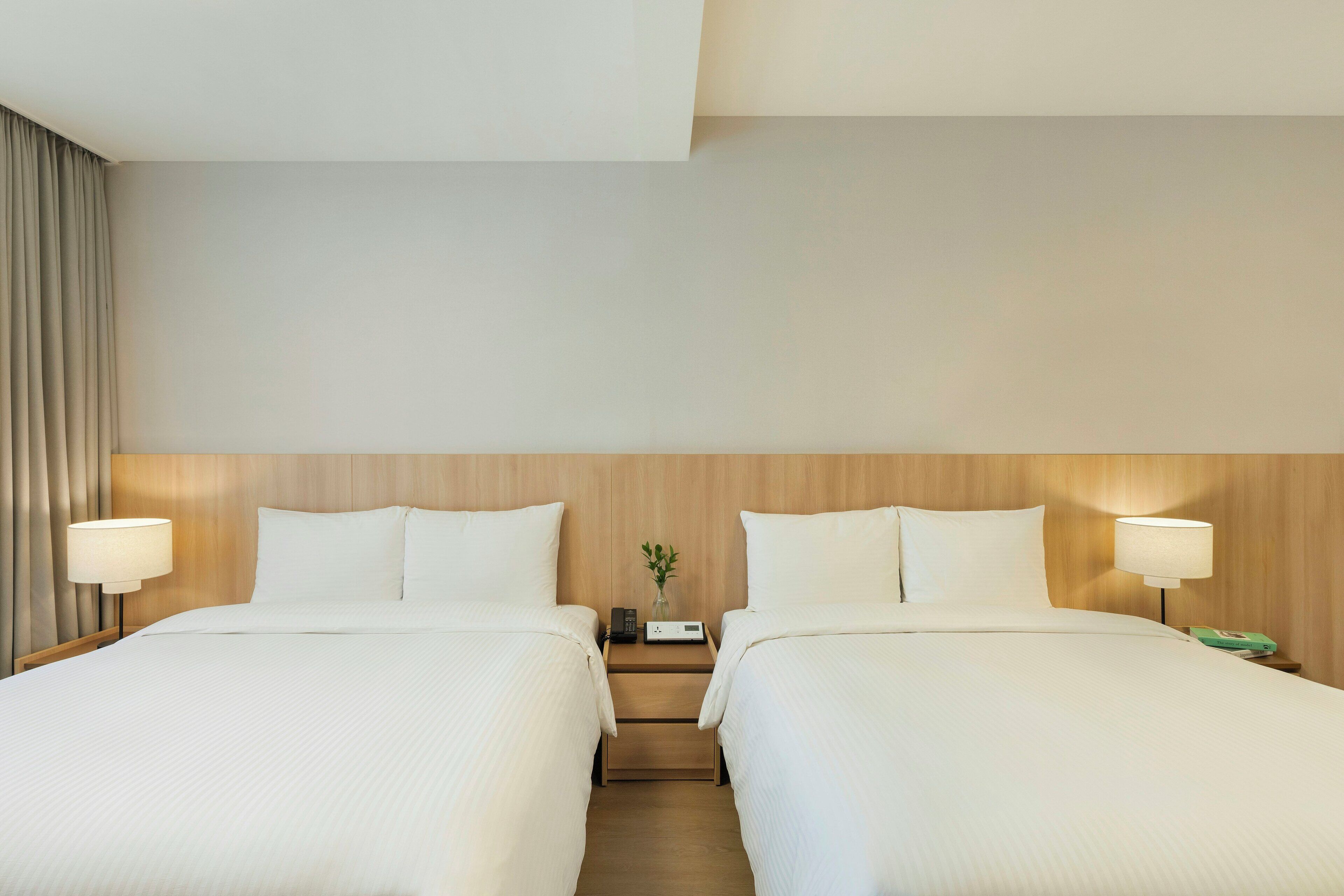 A hotel room with two beds, a wooden headboard, bedside lamps, a nightstand with a phone and a small vase with a plant, and a window with curtains.