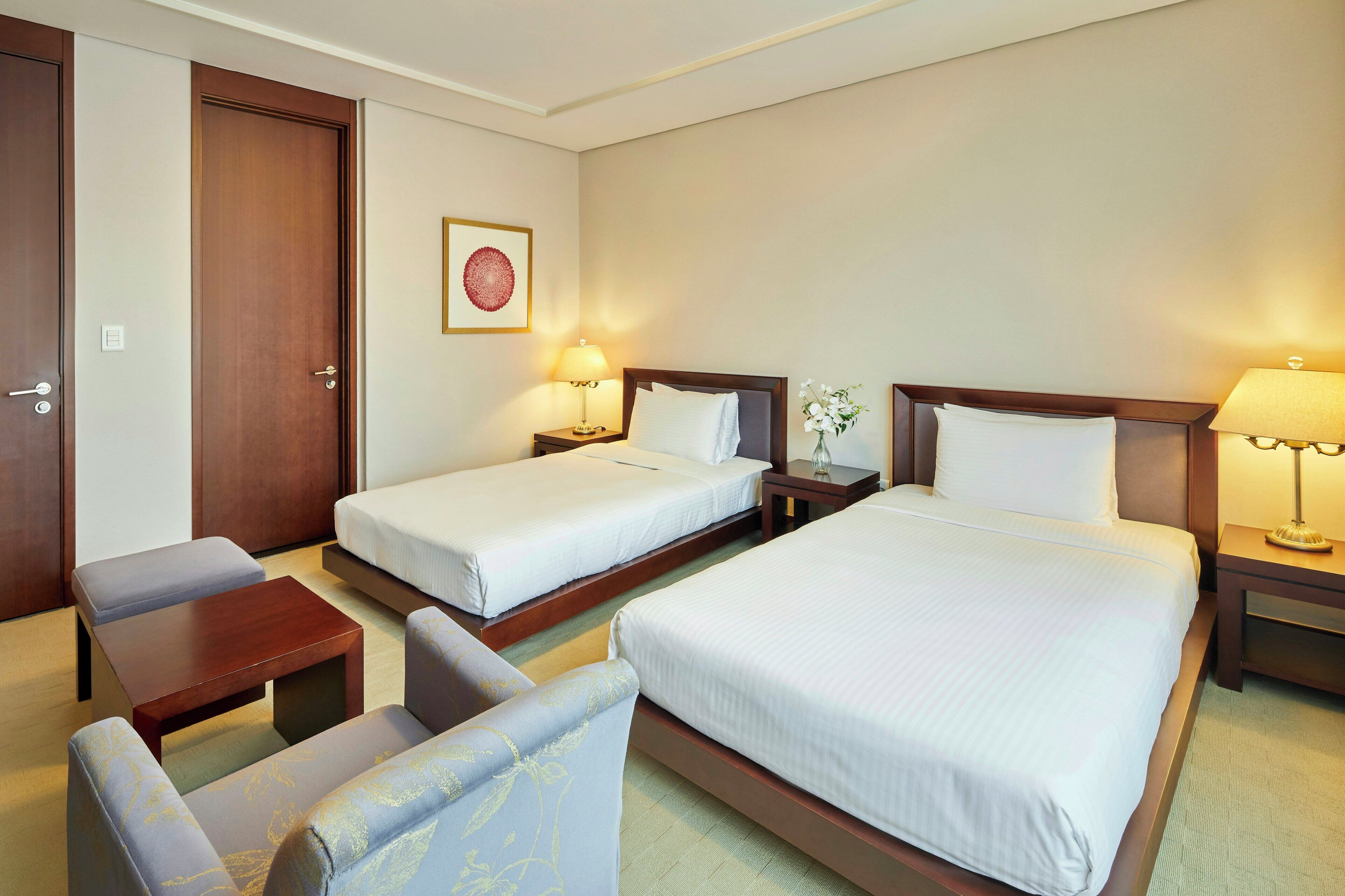 A hotel room with two single beds, a sofa, a small table, and a nightstand with a lamp.