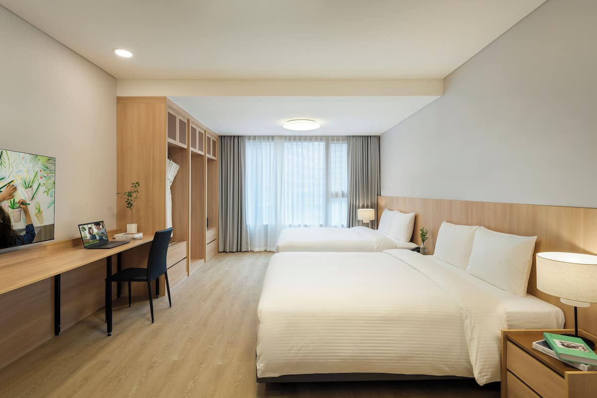 Habitación de hotel con escritorio, dos camas, una mesita de noche, lámpara y un cuadro en la pared.