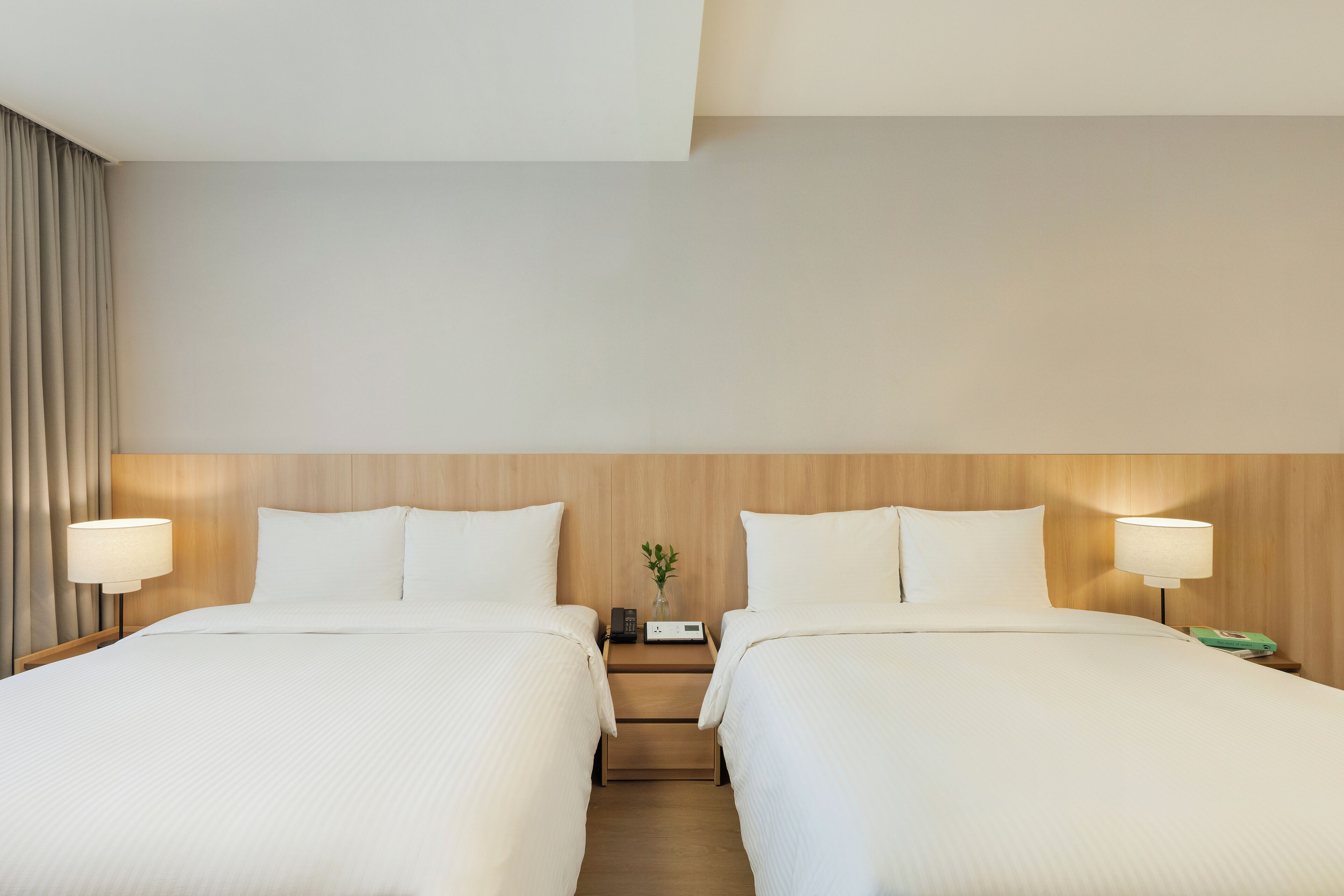 A hotel room with two beds, a wooden headboard, bedside lamps, a nightstand with a phone and a vase with a plant, and a curtain on the left side.