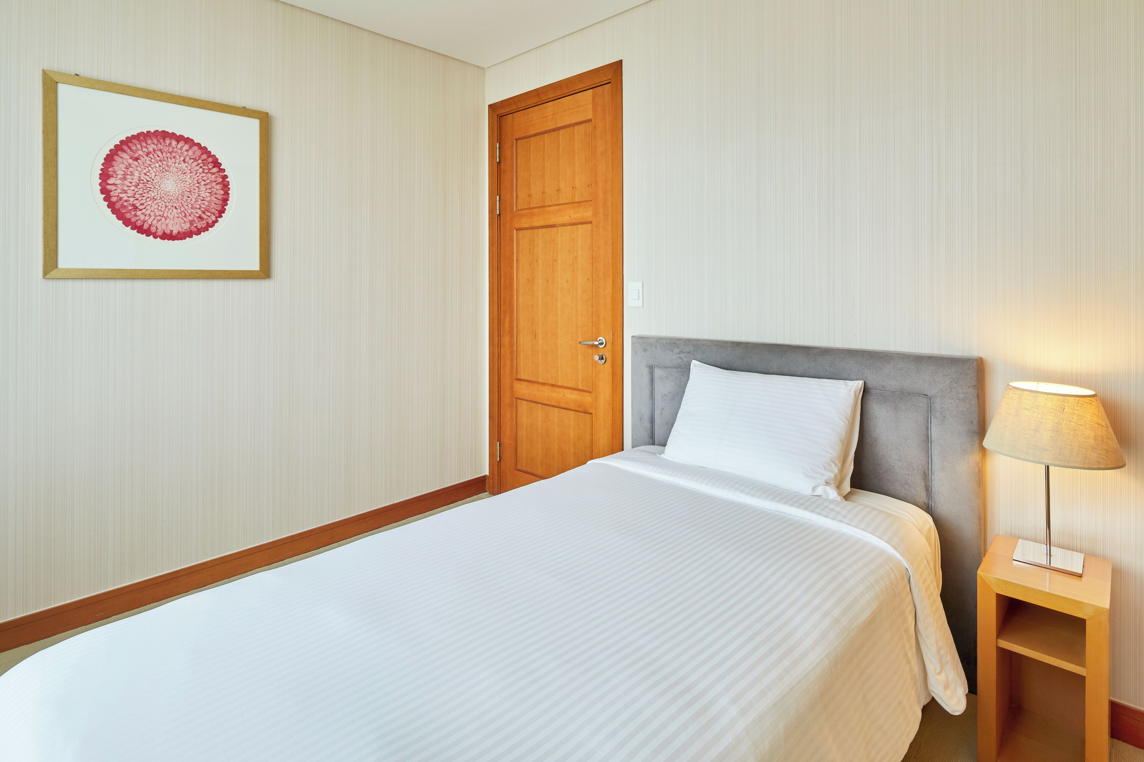 A neatly made bed with a grey headboard, a wooden door, and a framed artwork on the wall.