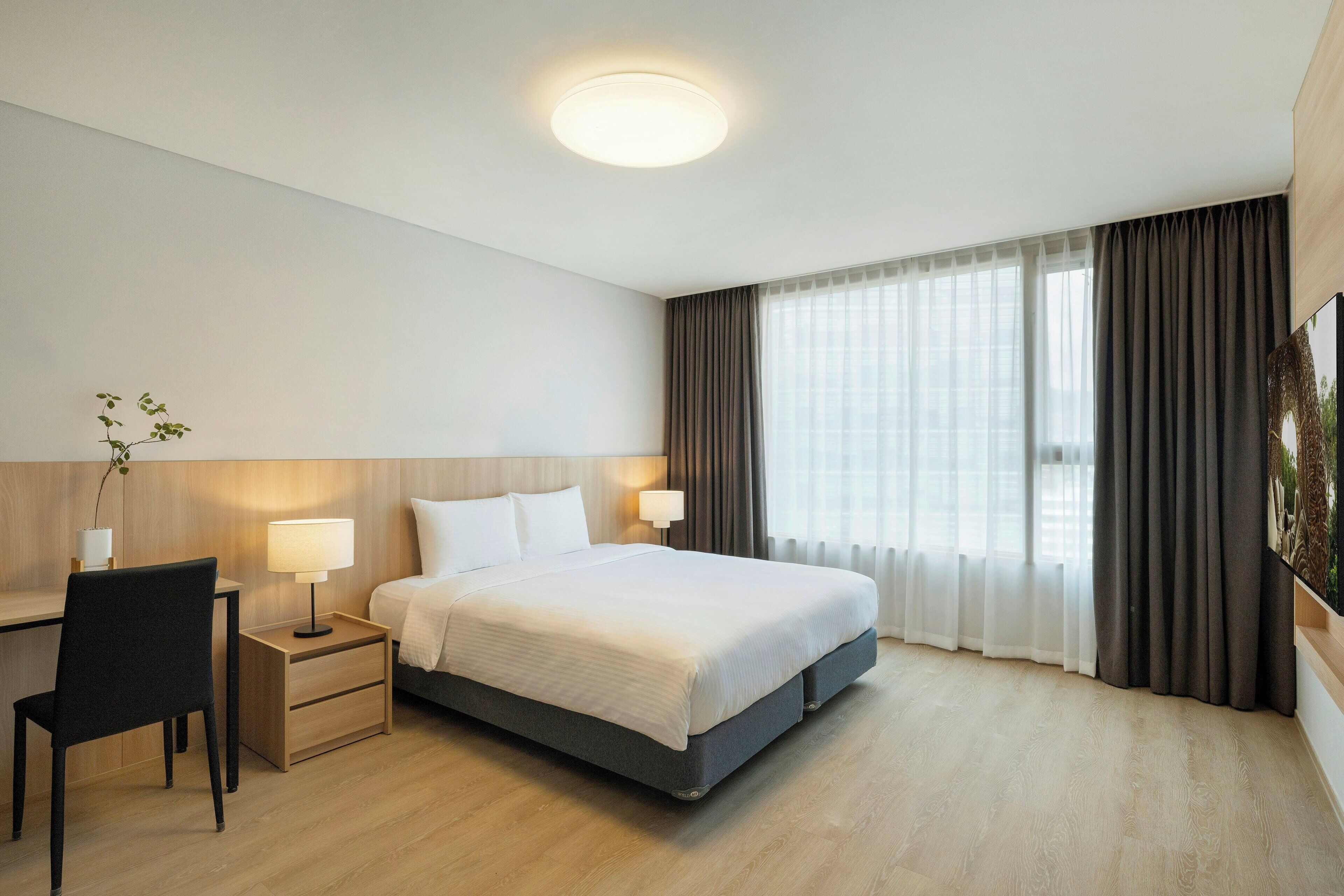 A hotel room with a bed, bedside tables, a desk, a chair, and a large window with curtains.
