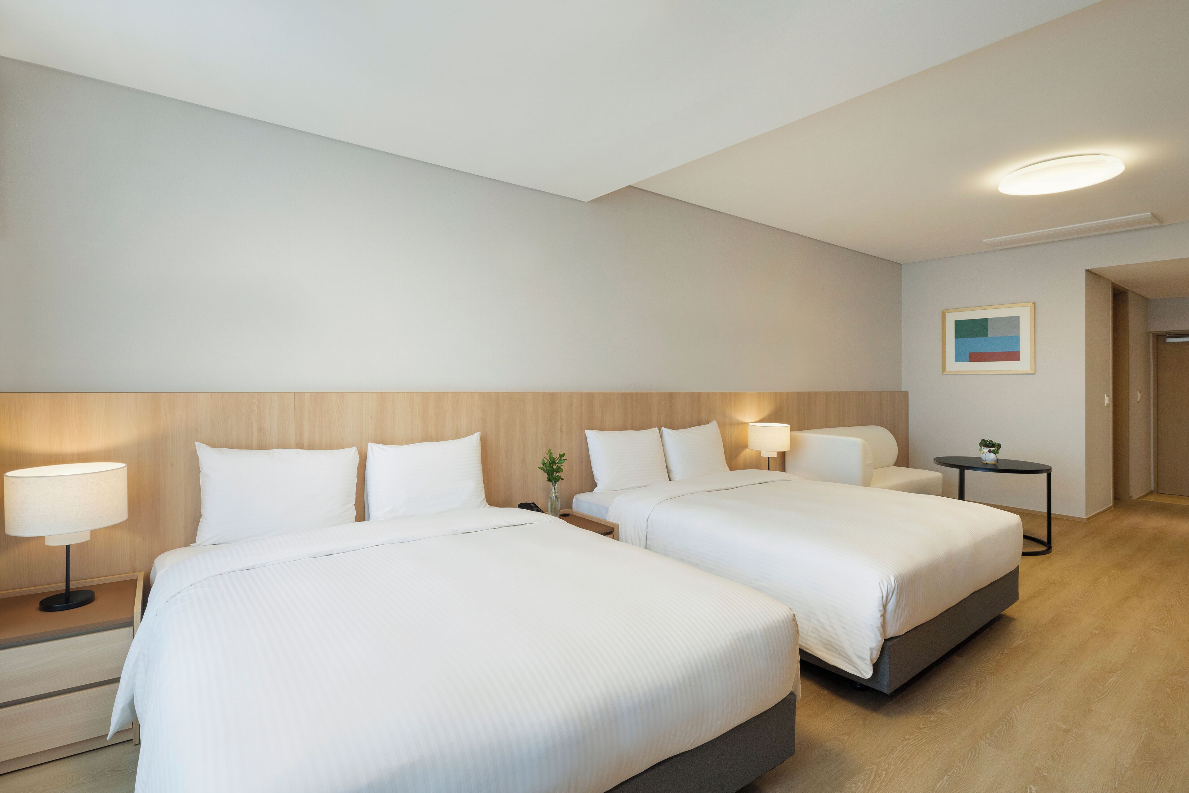 A hotel room with two beds, bedside tables, lamps, a small table with a plant, and a painting on the wall.