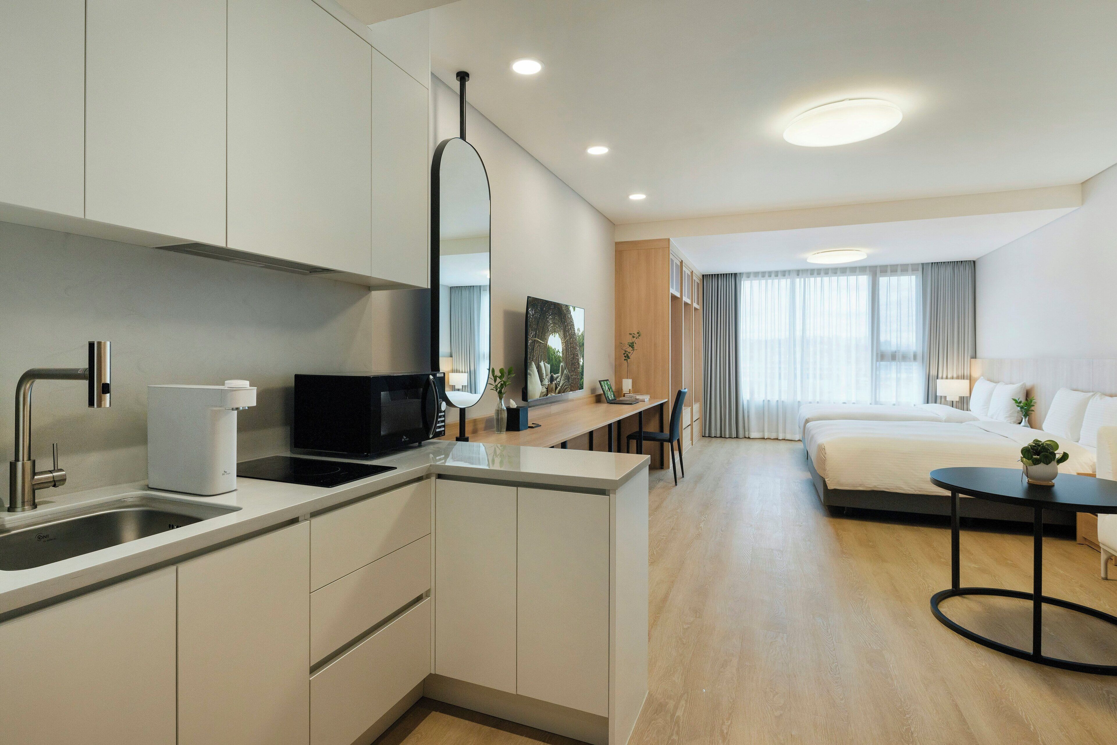 A modern hotel room with a kitchenette, a bed, a small table, and a view of the city.