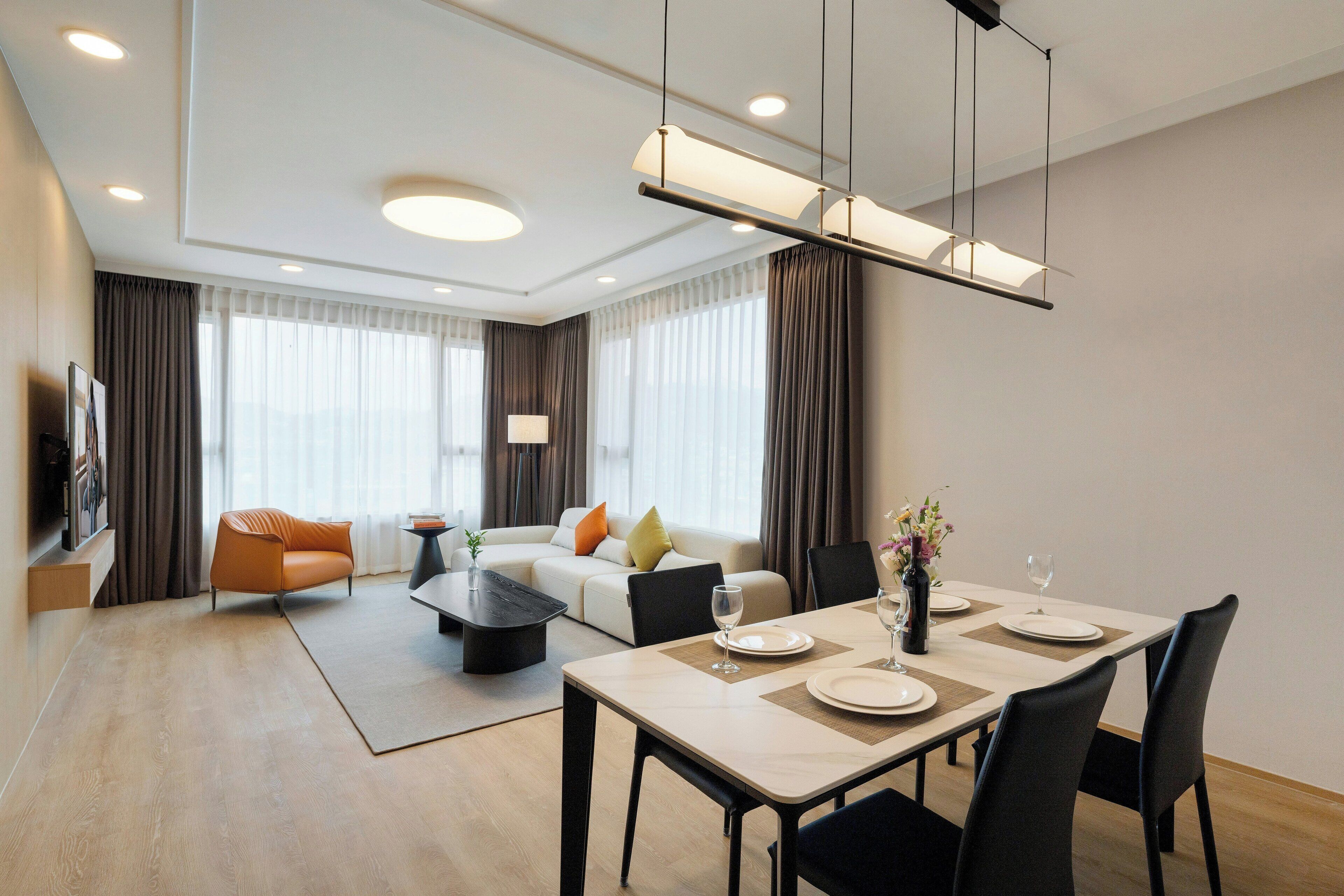 Una habitación de hotel moderna con zona de comedor, sofá, televisión y lámpara.