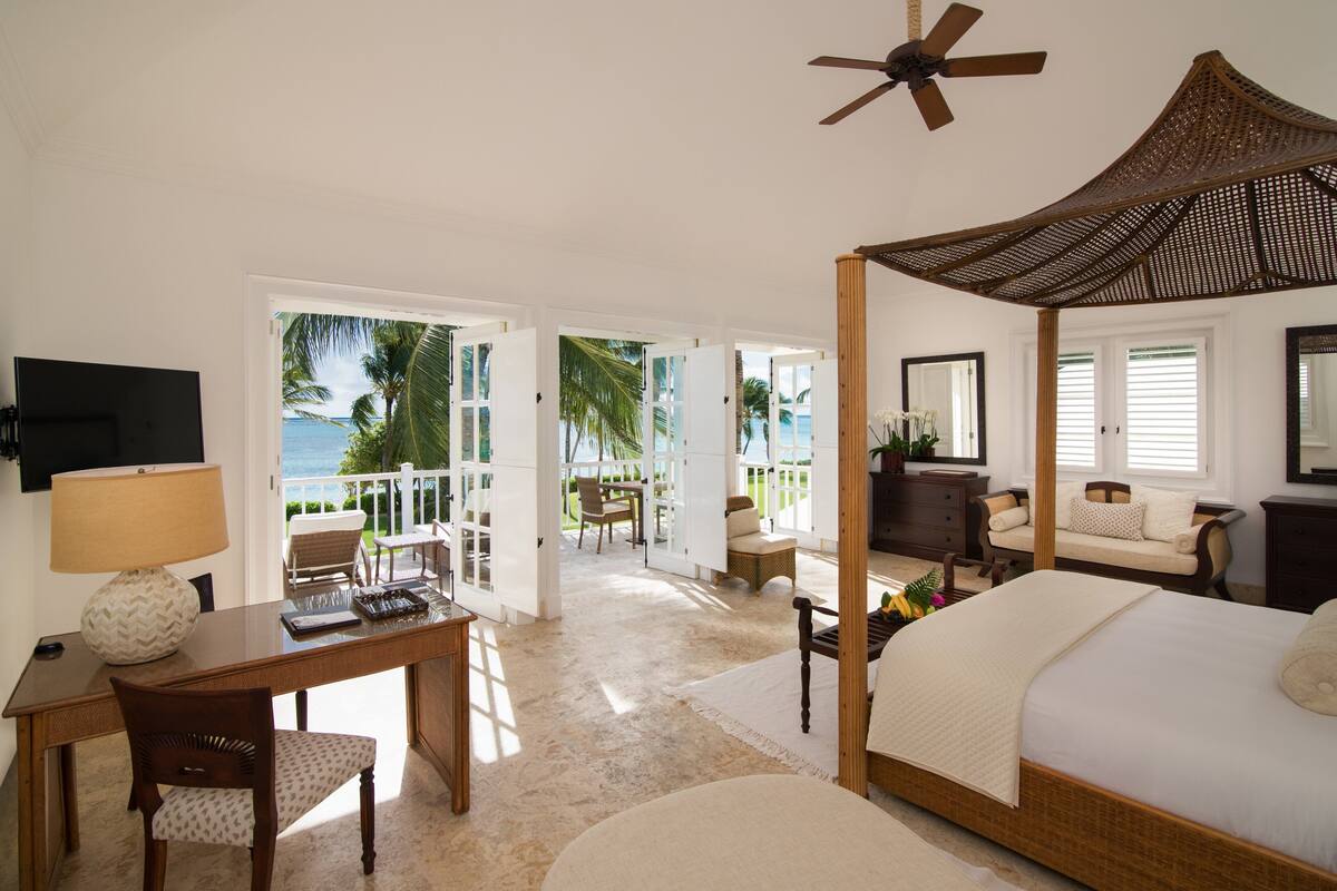 A bedroom with a four-poster bed, a ceiling fan, and a view of the ocean through open doors.