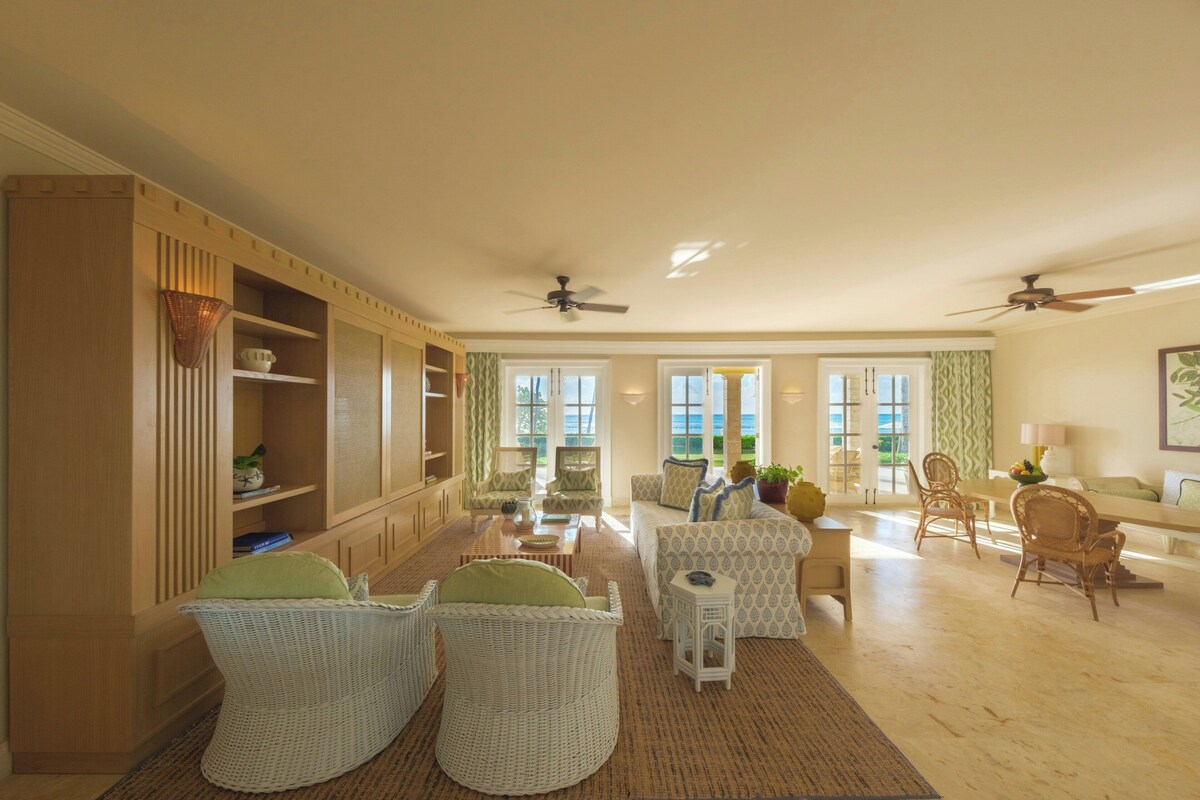 A spacious living room with a large window, wicker furniture, and a ceiling fan.