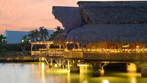 A thatched-roof overwater bungalow with a wooden deck and string lights, overlooking calm water. - Tortuga Bay Hotel (Punta Cana)