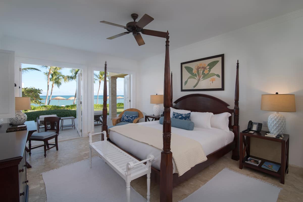 A four-poster bed with a view of the ocean, a ceiling fan, and a painting of flowers on the wall.