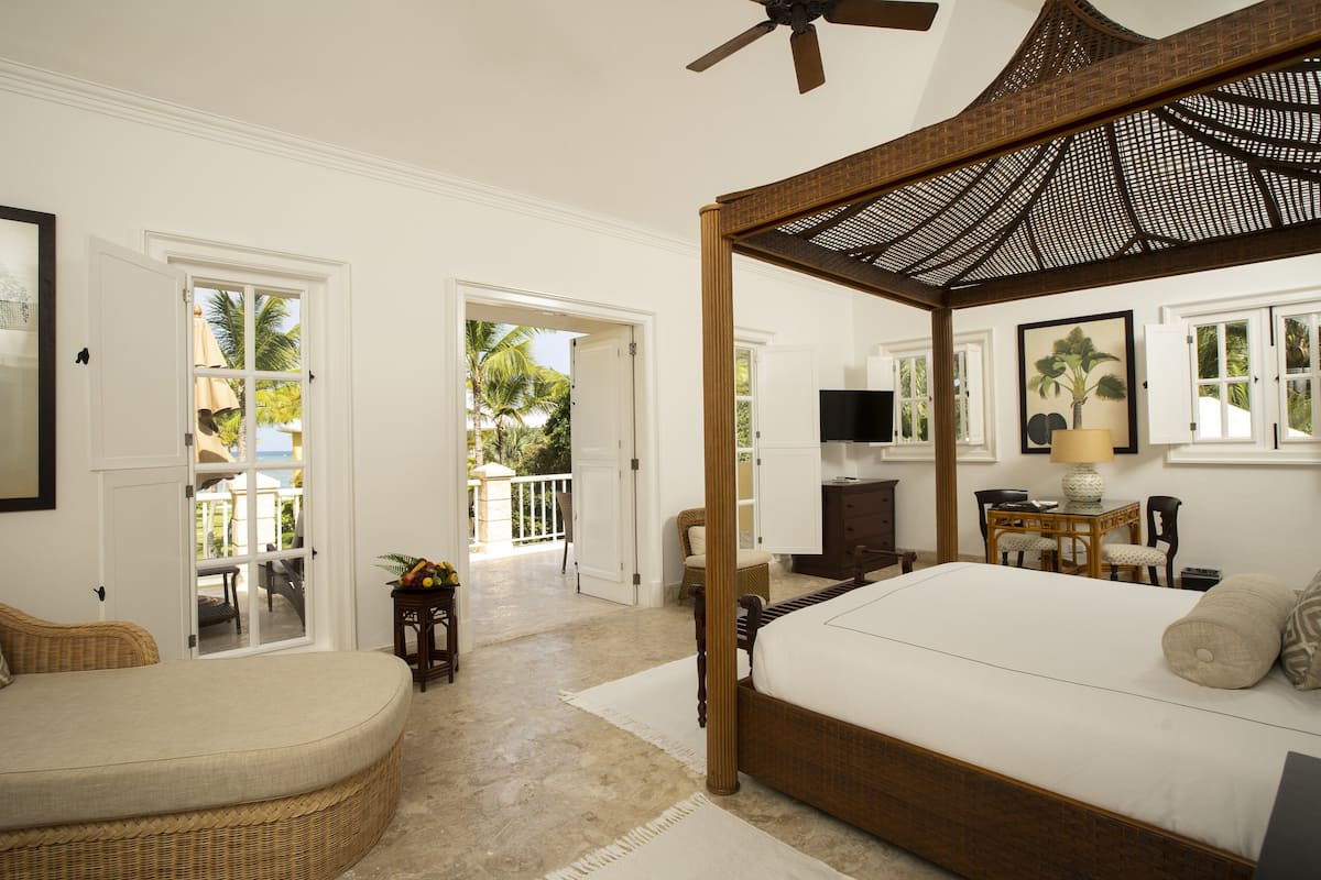 A bedroom with a four-poster bed, a television, a desk with a lamp, and a view of the outdoors through an open door.