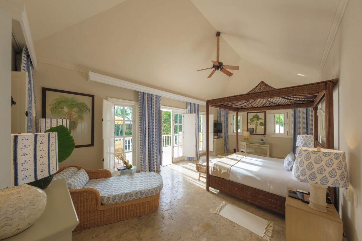 A spacious bedroom with a four-poster bed, a sitting area with a wicker sofa, and a large window providing a view of the outdoors.
