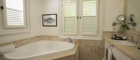 A bathroom with a large bathtub, a sink, and a vanity with a mirror. There are two windows with blinds, and a framed picture on the wall.