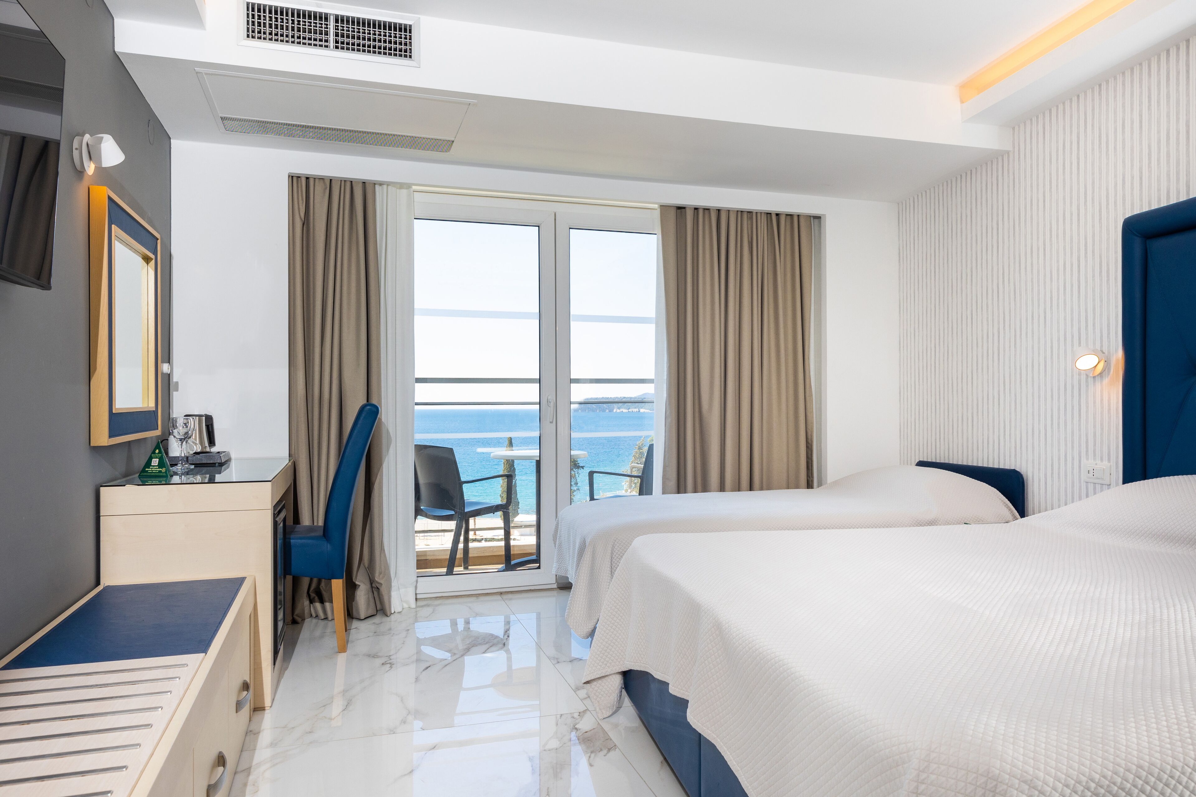 Ein Hotelzimmer mit zwei Betten, einem Schreibtisch, einem Stuhl und einem Balkon mit Meerblick.