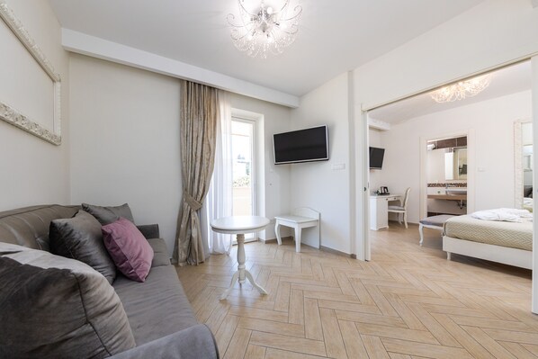 Una sala moderna con un sofá, una mesita redonda, un televisor montado en la pared y un dormitorio con cama y escritorio.