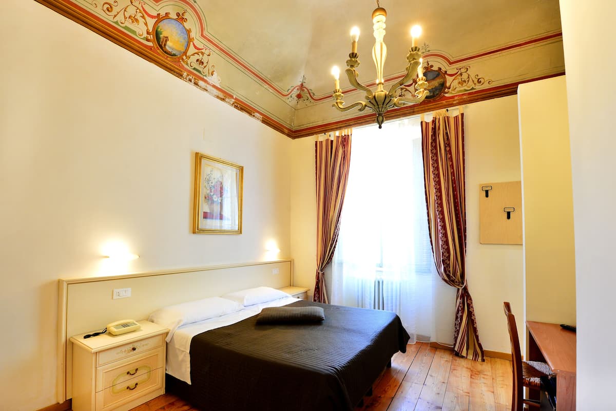A hotel room with a bed, bedside table, lamp, curtains, and a chandelier.
