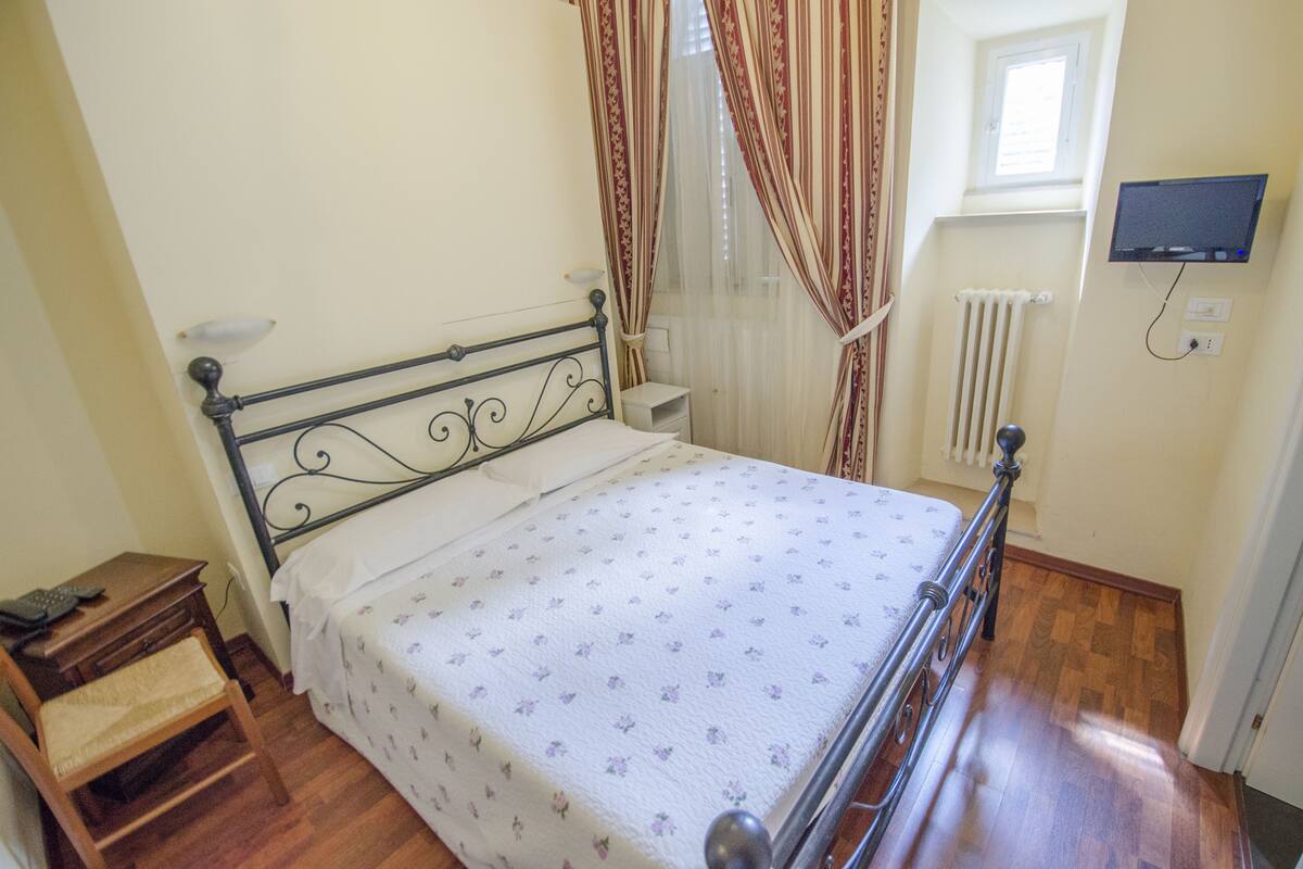 A bedroom with a bed, wooden floor, curtains, a bedside table, a chair, a TV on the wall, and a window with a radiator.