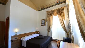 Une chambre d’hôtel avec une tête de lit en bois, un lit recouvert d’une couverture noire, une table de chevet avec une lampe et une fenêtre avec des rideaux.
