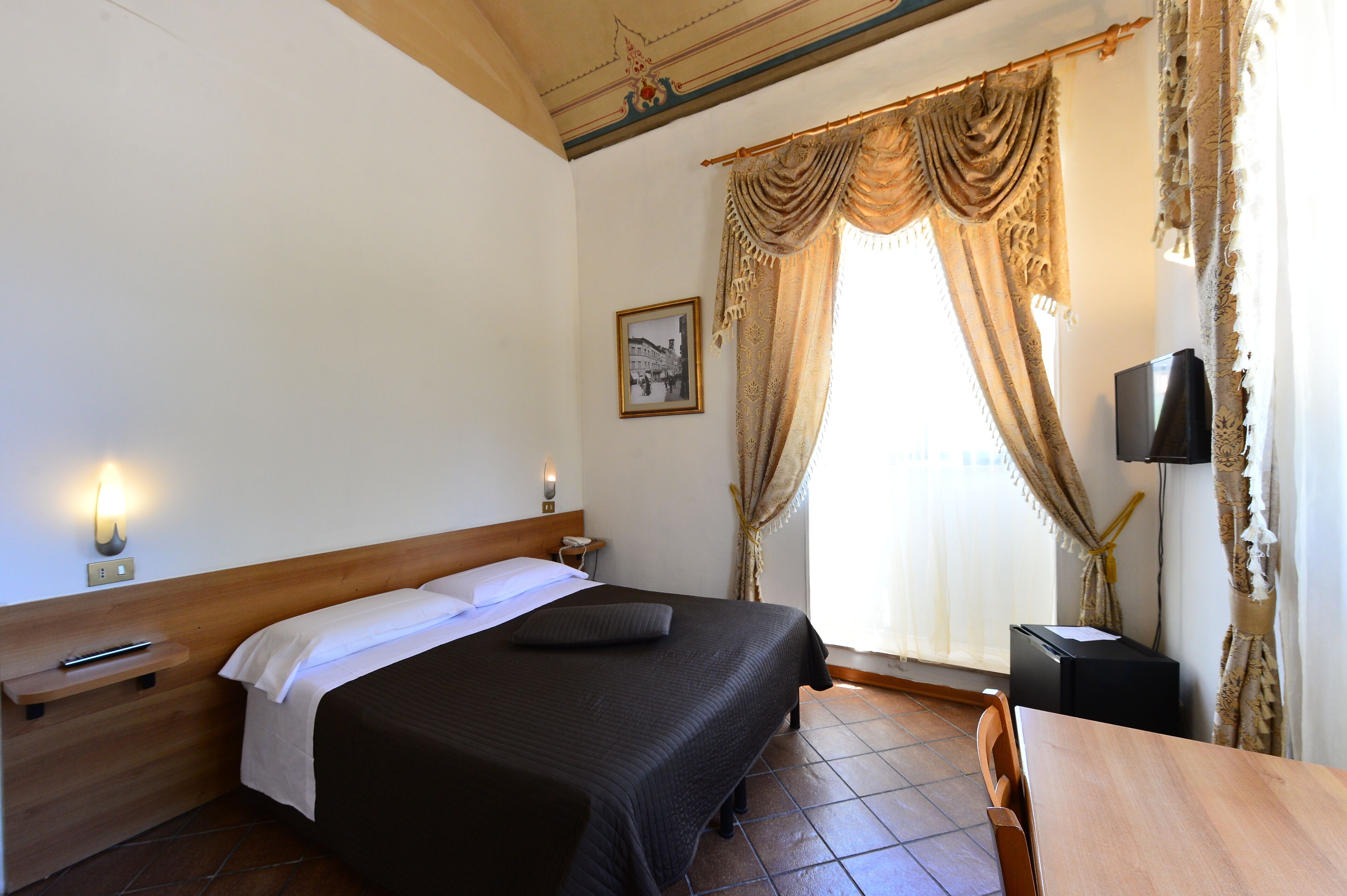 Una camera d'albergo con un letto, comodino, lampada, televisore e tende.