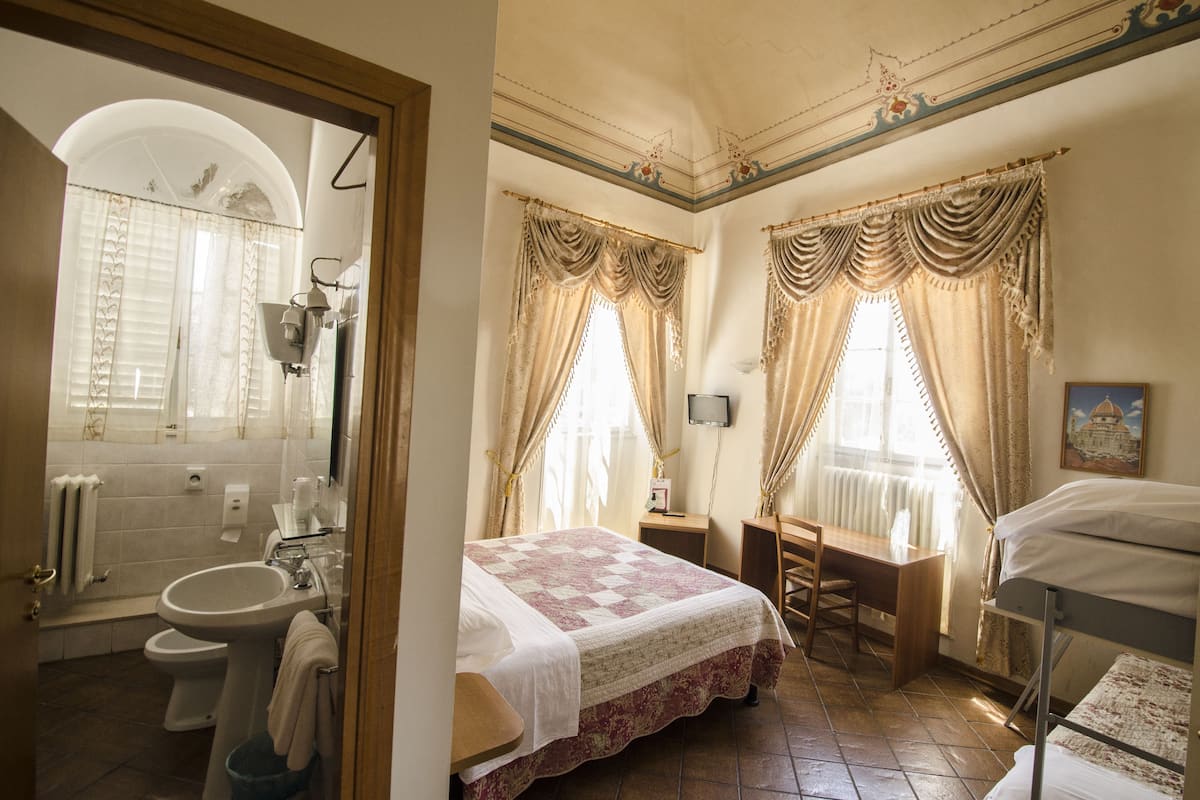 A bedroom with a bed, a desk, a chair, a window with curtains, a radiator, and a bathroom with a sink and toilet.