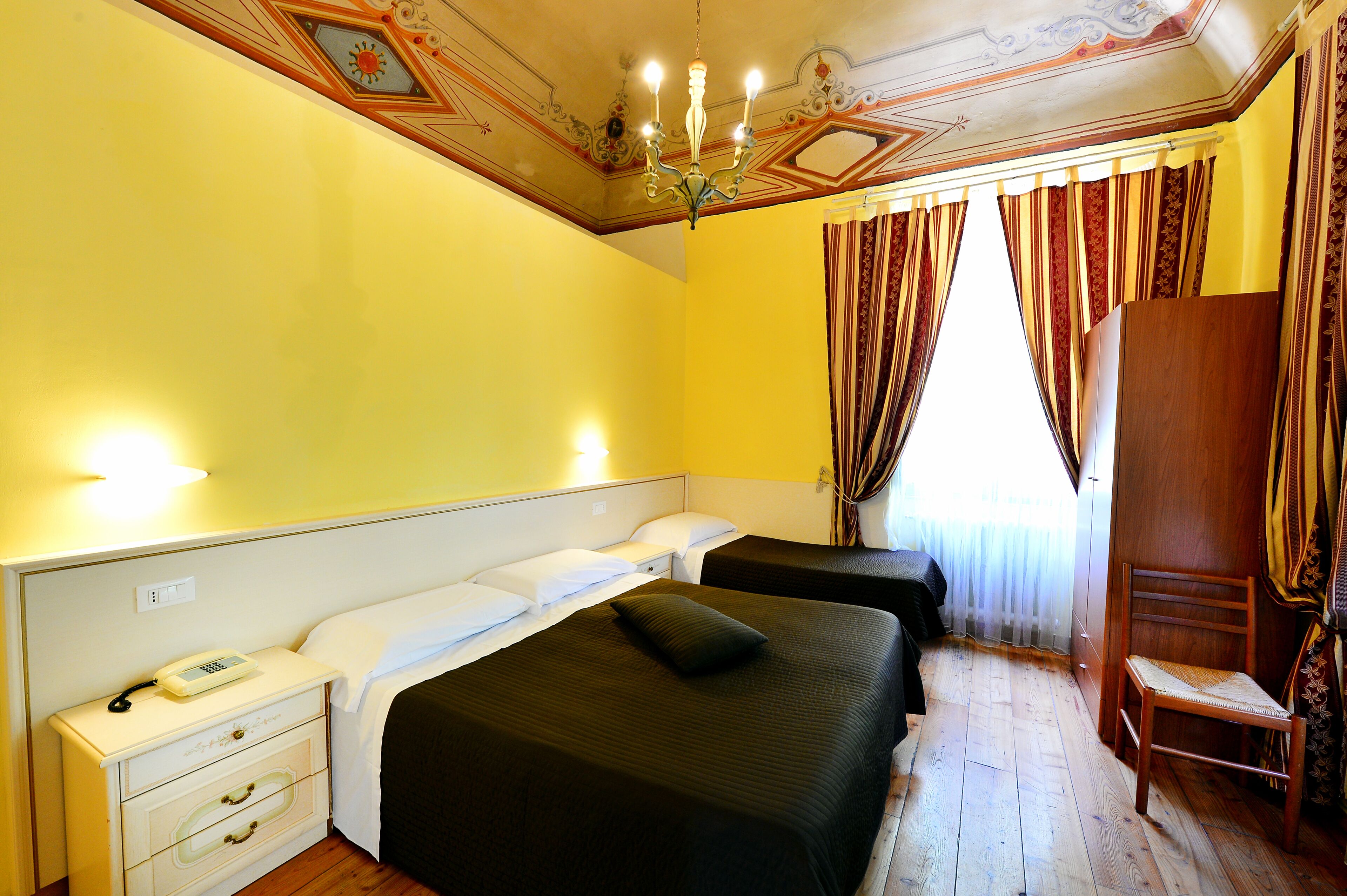 Habitación de hotel con dos camas, una silla de madera, una mesita de noche con un teléfono y un techo decorativo.