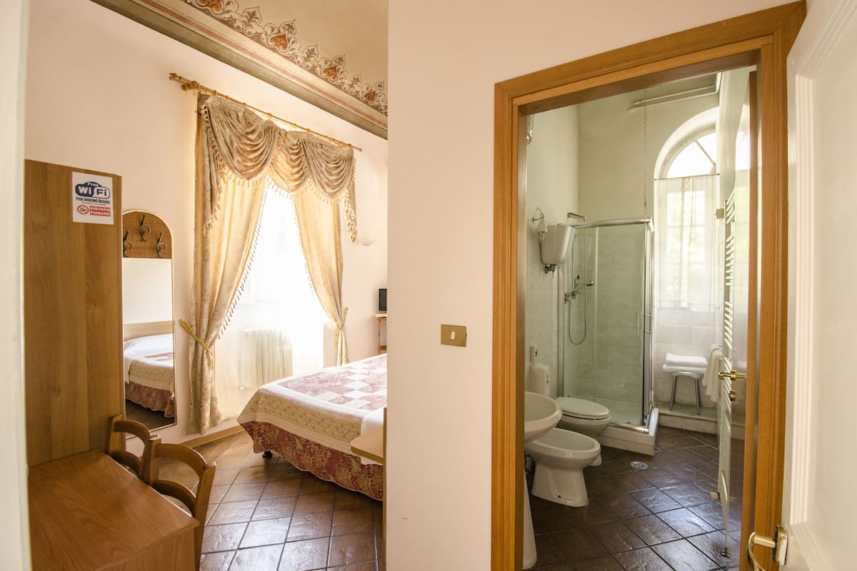 A bedroom with a bed, a wooden chair, a bedside table, and a window with curtains. The bathroom includes a toilet, a shower, and a glass-enclosed shower area.