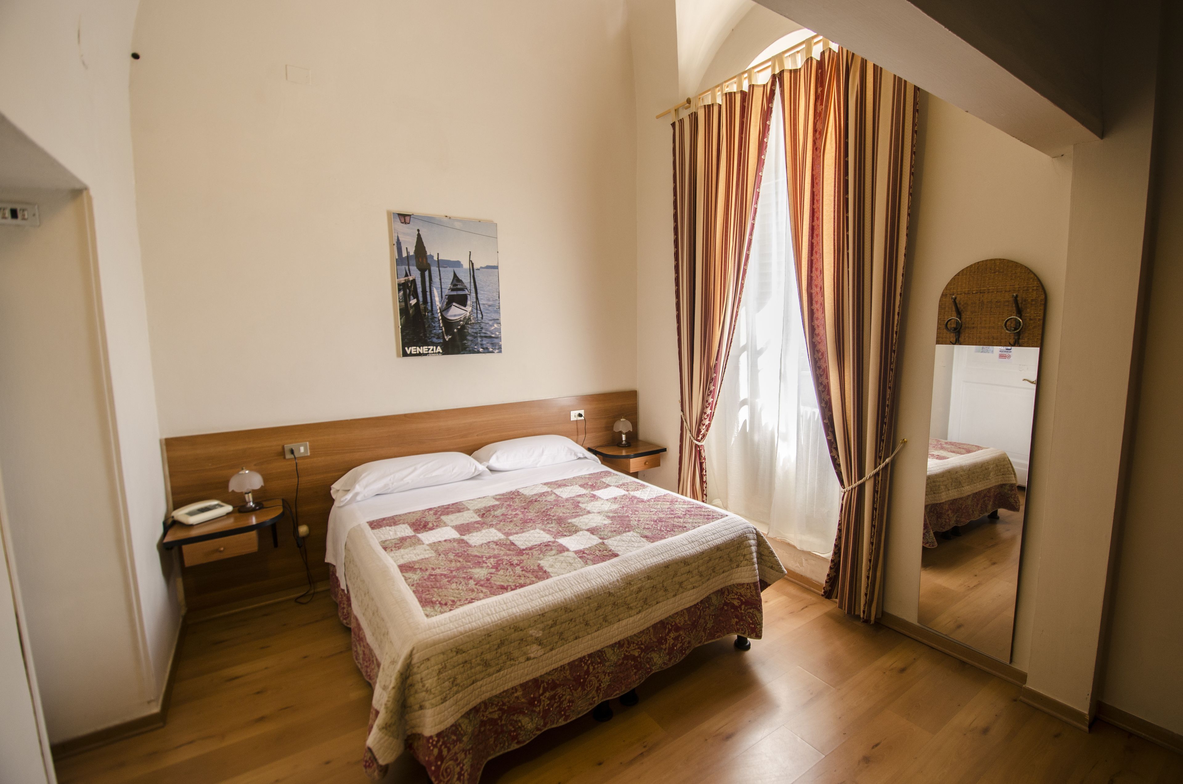A bedroom with a wooden headboard, a bed with a patterned bedspread, a nightstand with a lamp, a wall-mounted picture, and a window with curtains.