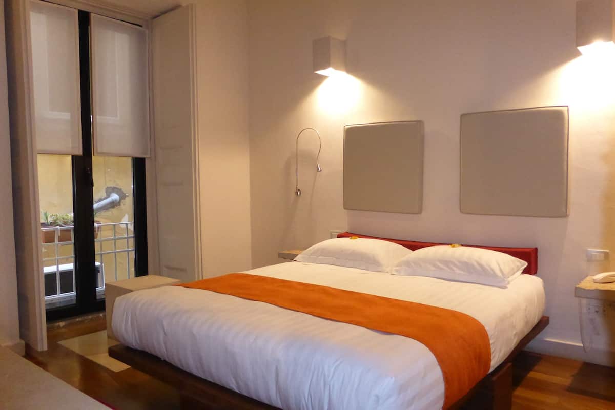 A bedroom with a bed, a nightstand, a lamp, and a window with a view of the outside.