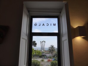 A view through a window with the word "MICALDO" on it, overlooking a landscaped area with palm trees and a distant view of the sea. - Micalò ArtRooms (Naples)