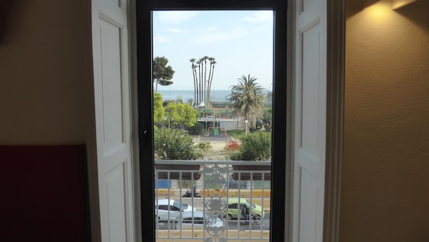 A view through a door with a metal gate, leading to a parking area with cars and palm trees. - Micalò ArtRooms (Naples)