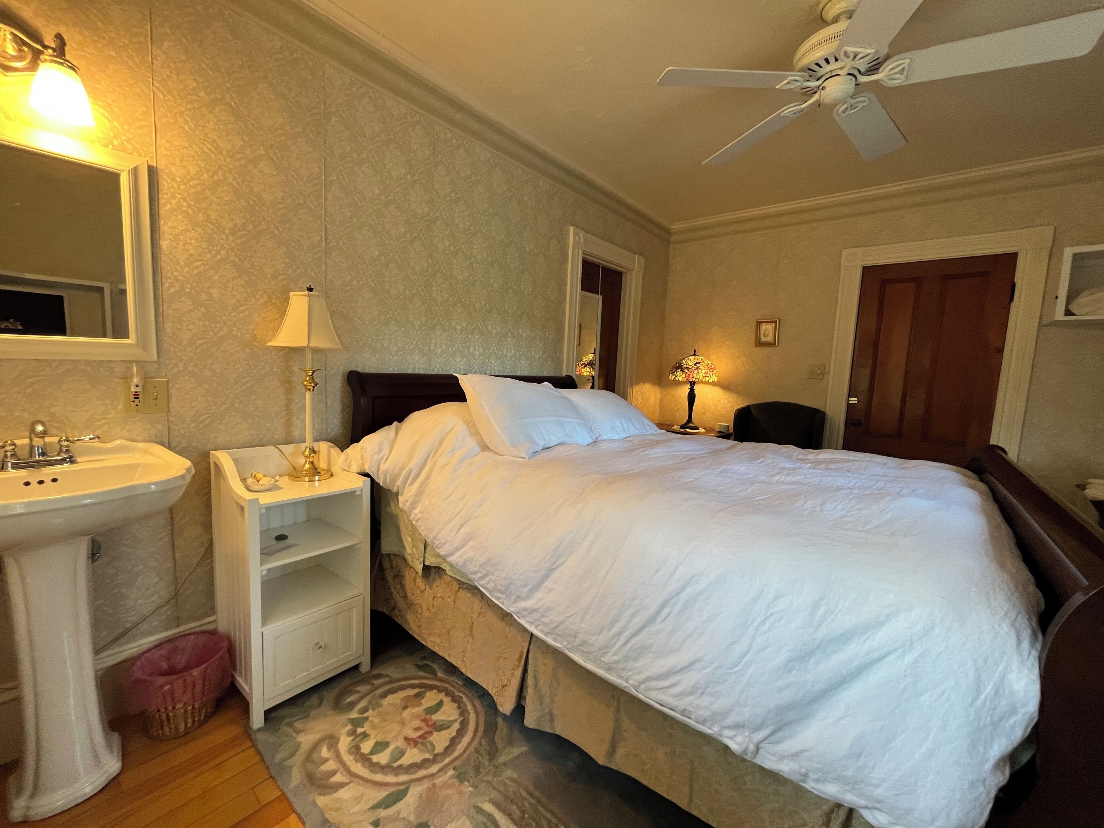 Ein Hotelzimmer mit einem Bett, einem Nachttisch mit Lampe, einem Spiegel, einem Deckenventilator und einer Tür.