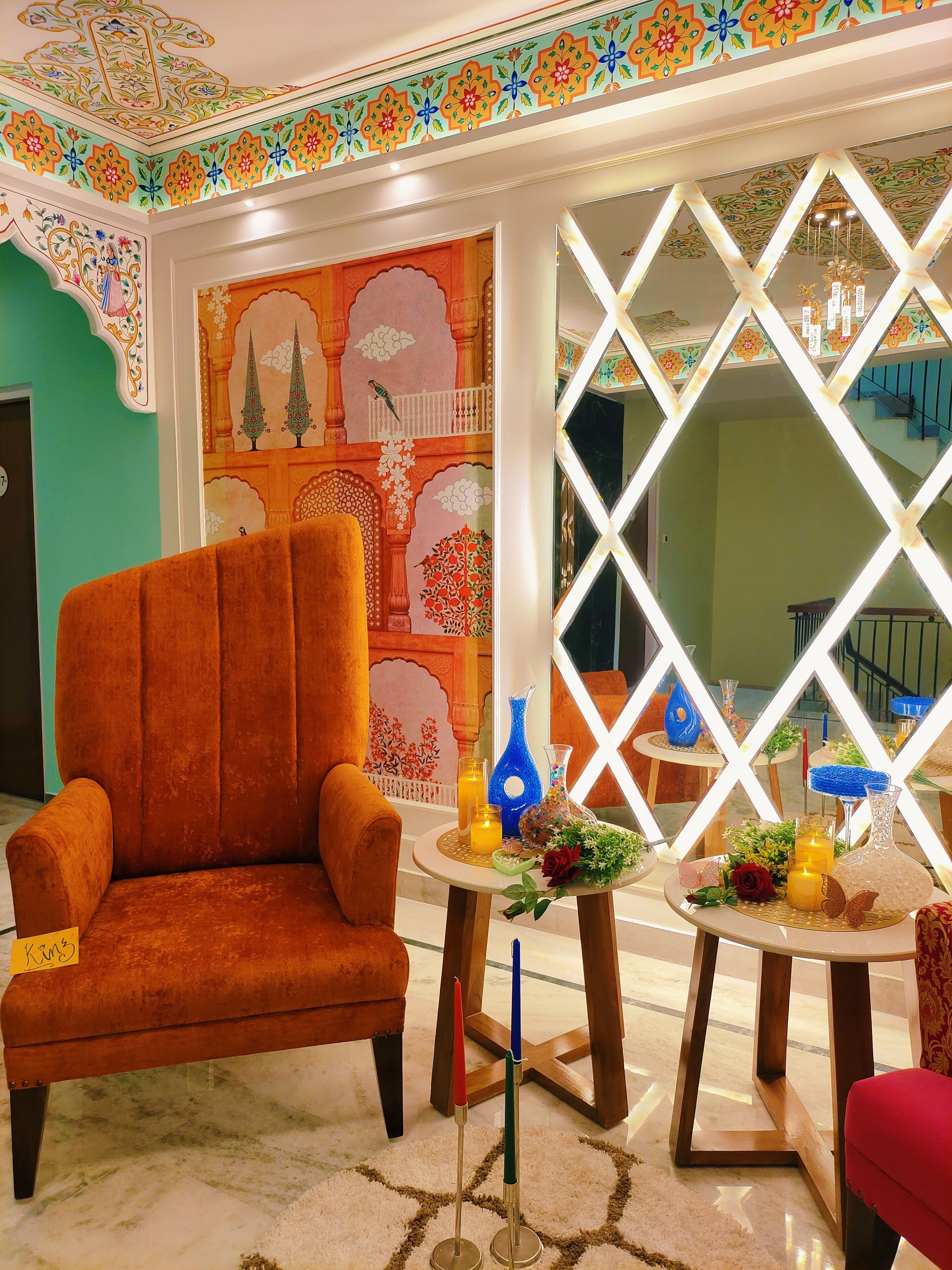 A room with a plush orange armchair, a decorative wall with floral patterns, and a table with candles and vases.