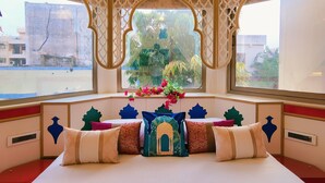 Una habitación con una cama grande, almohadillas decorativas, una araña y un ventanal con vistas a edificios y vegetación.