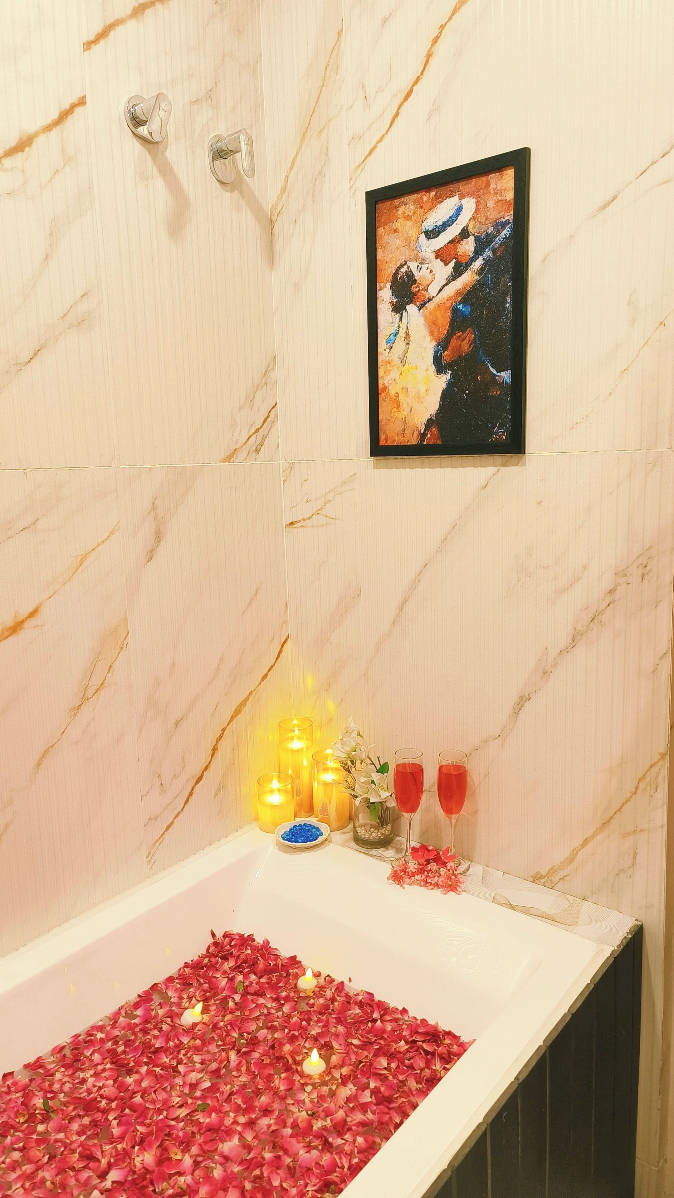 A bathroom with a marble wall, a framed painting, candles, a glass jar with beads, and a bathtub filled with rose petals.