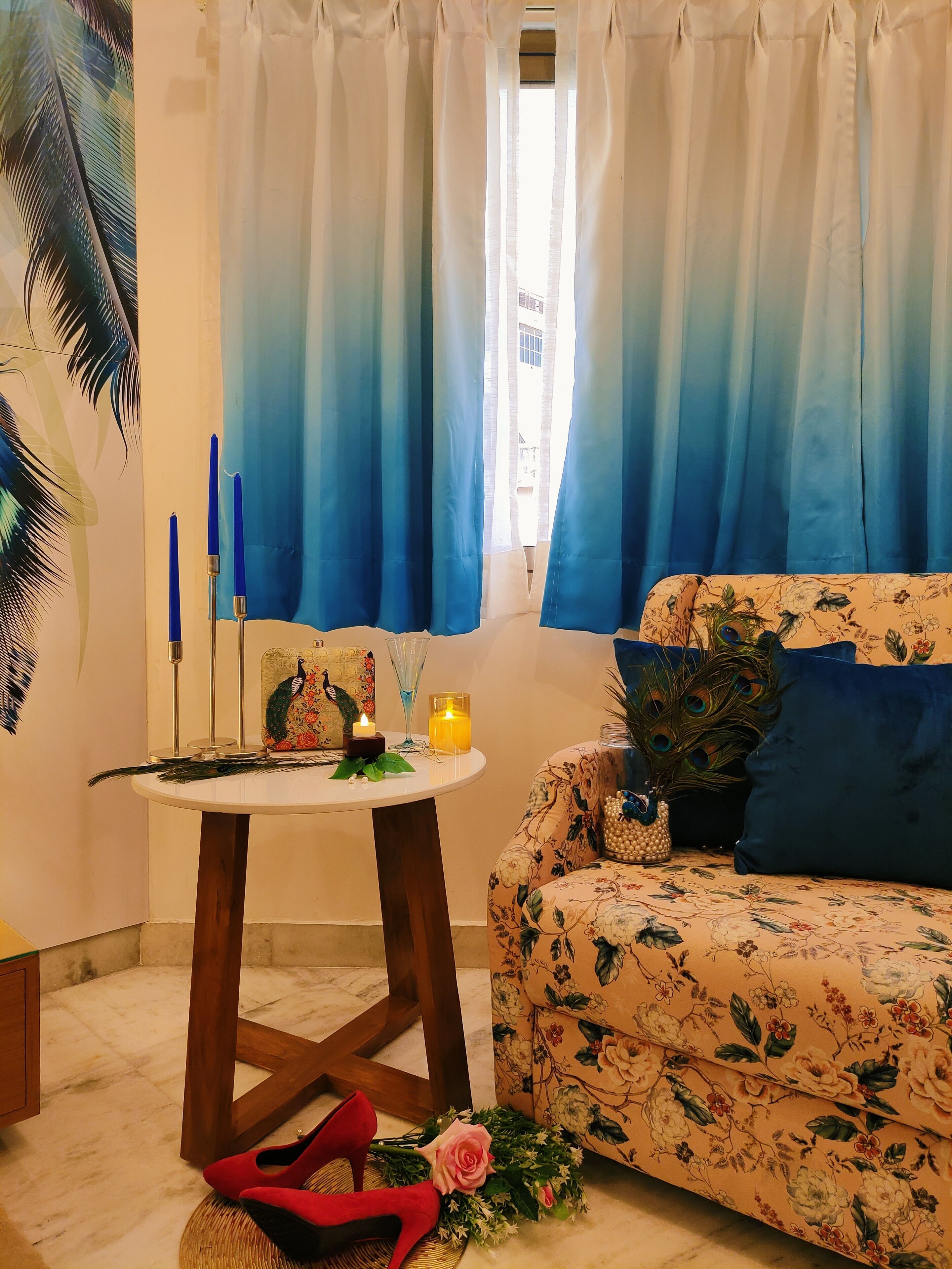 Een kamer met een bloemenbank, een ronde tafel met een kaars, een decoratie van een pauw en een raam met blauwe gordijnen.