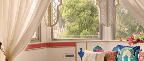 Quarto com uma cama grande, janela com vista para vegetação e um banquinho vermelho com chinelos.