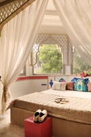 Quarto com uma cama grande, janela com vista para vegetação e um banquinho vermelho com chinelos.