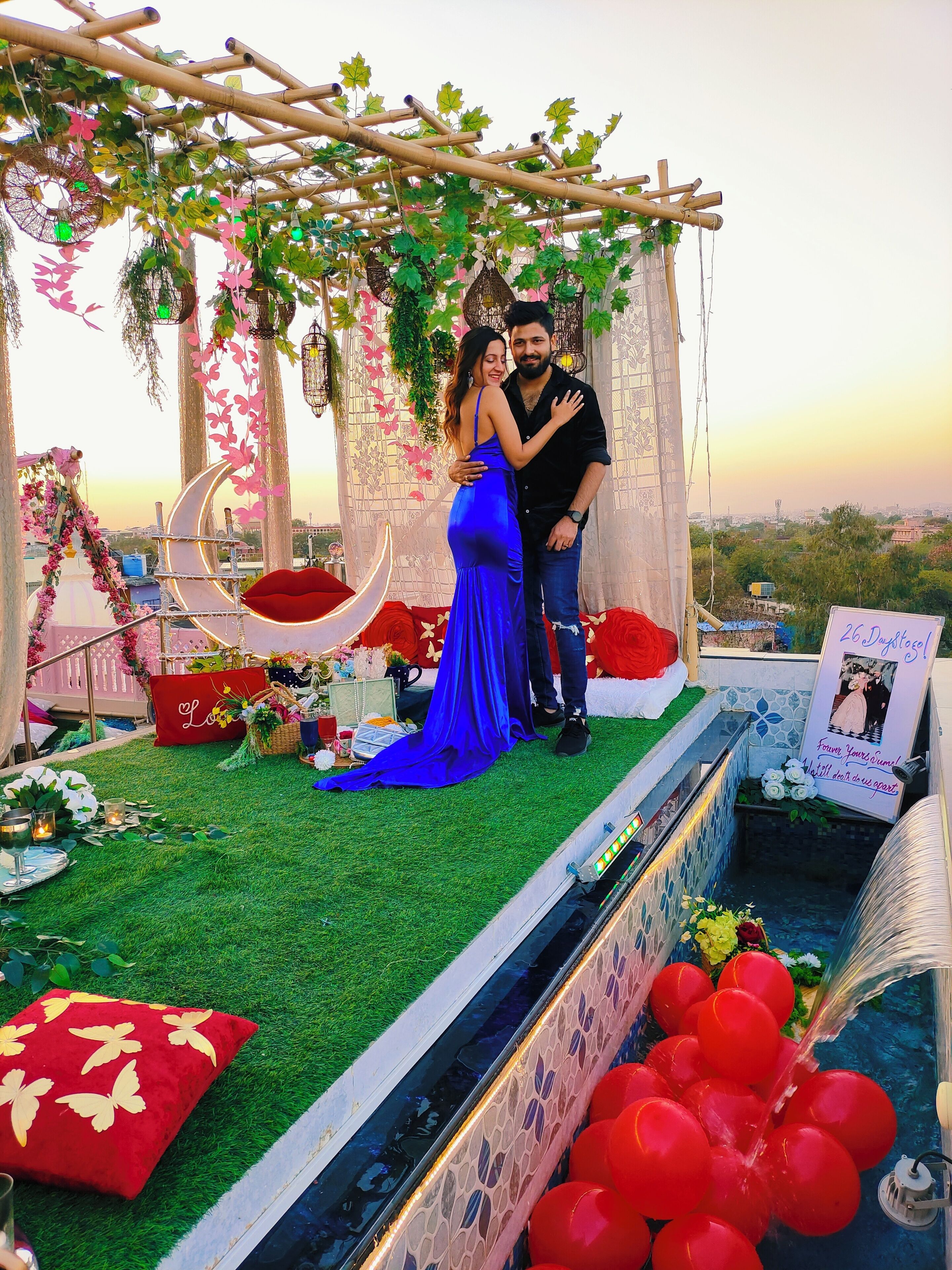 Una pareja de pie en un escenario exterior decorado con fondo de bambú y follaje, una pequeña piscina con globos rojos y un cartel con una foto y texto.