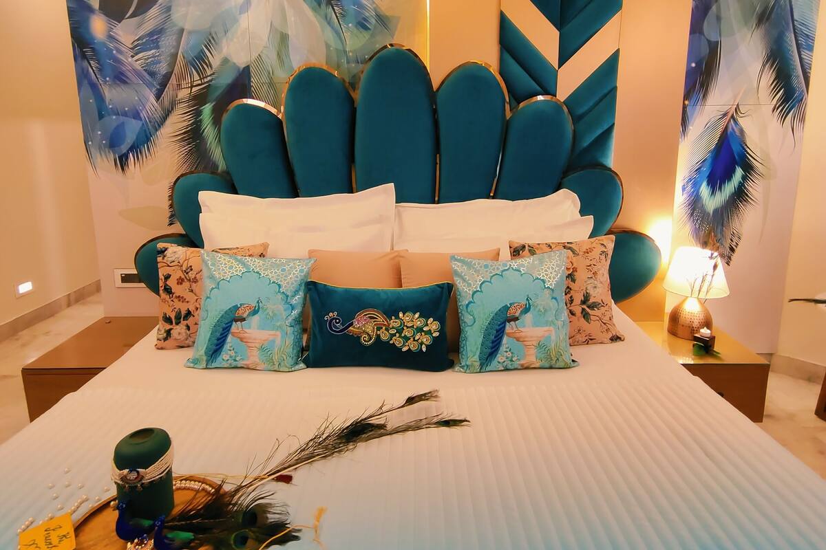 Une chambre à coucher avec un grand lit, une tête de lit recouverte d’un tissu turquoise, des coussins décoratifs et un mur orné d’une fresque représentant des plumes de paon.