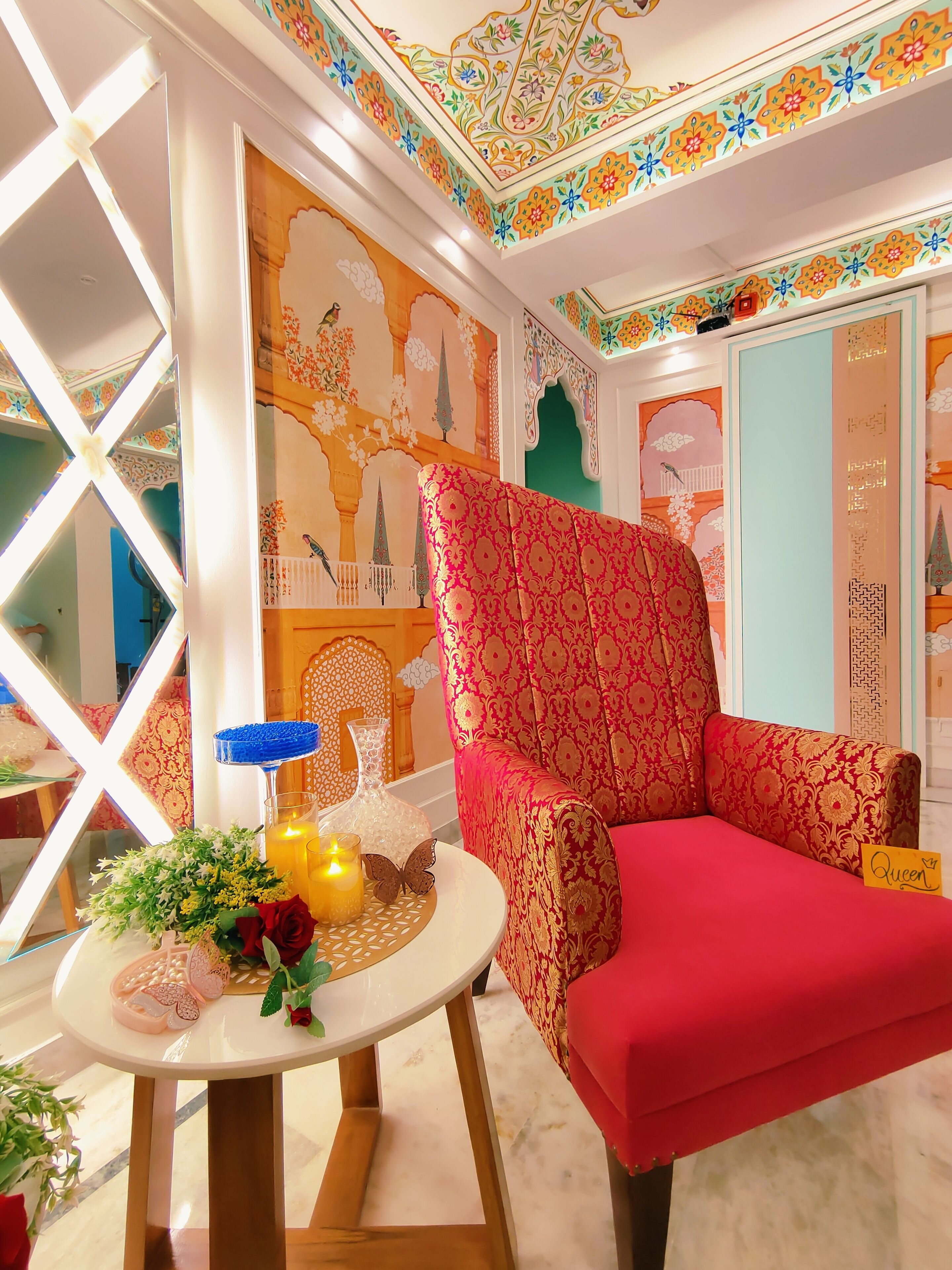 A room with a red upholstered chair, a small round table with a candle, and a vase with flowers. The walls have intricate floral and geometric patterns.