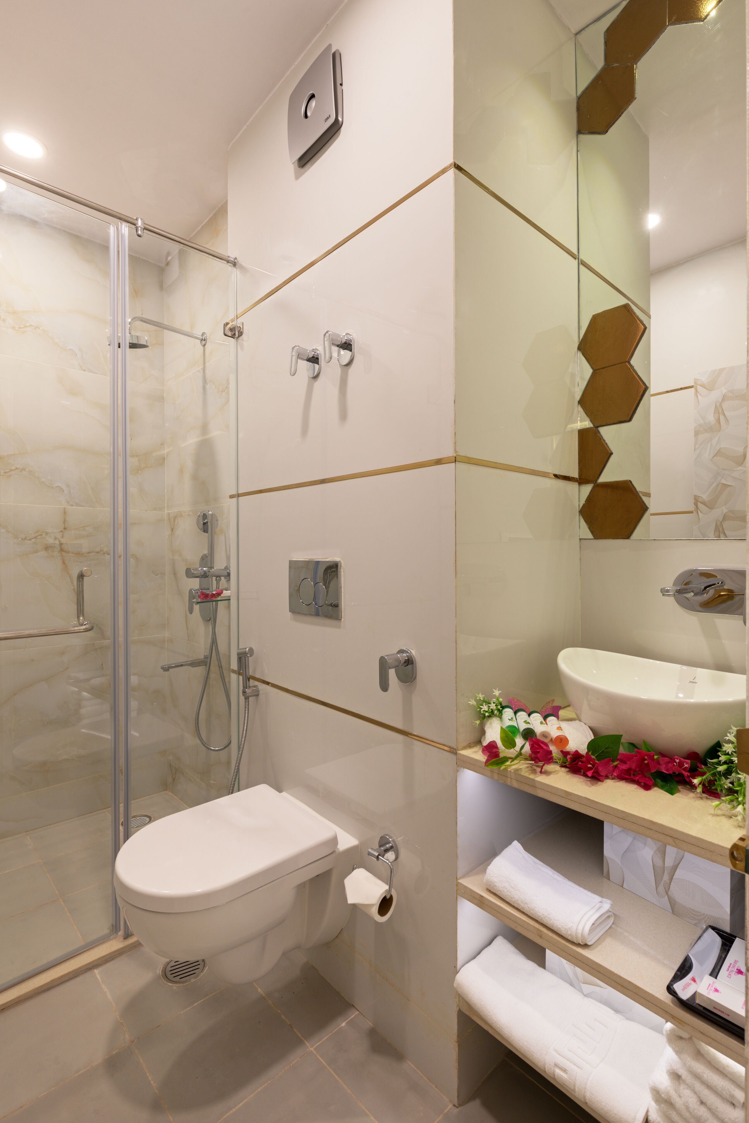 Un baño moderno con cabina de ducha de vidrio, inodoro blanco, lavamanos con espejo y estante con toallas y artículos de aseo.